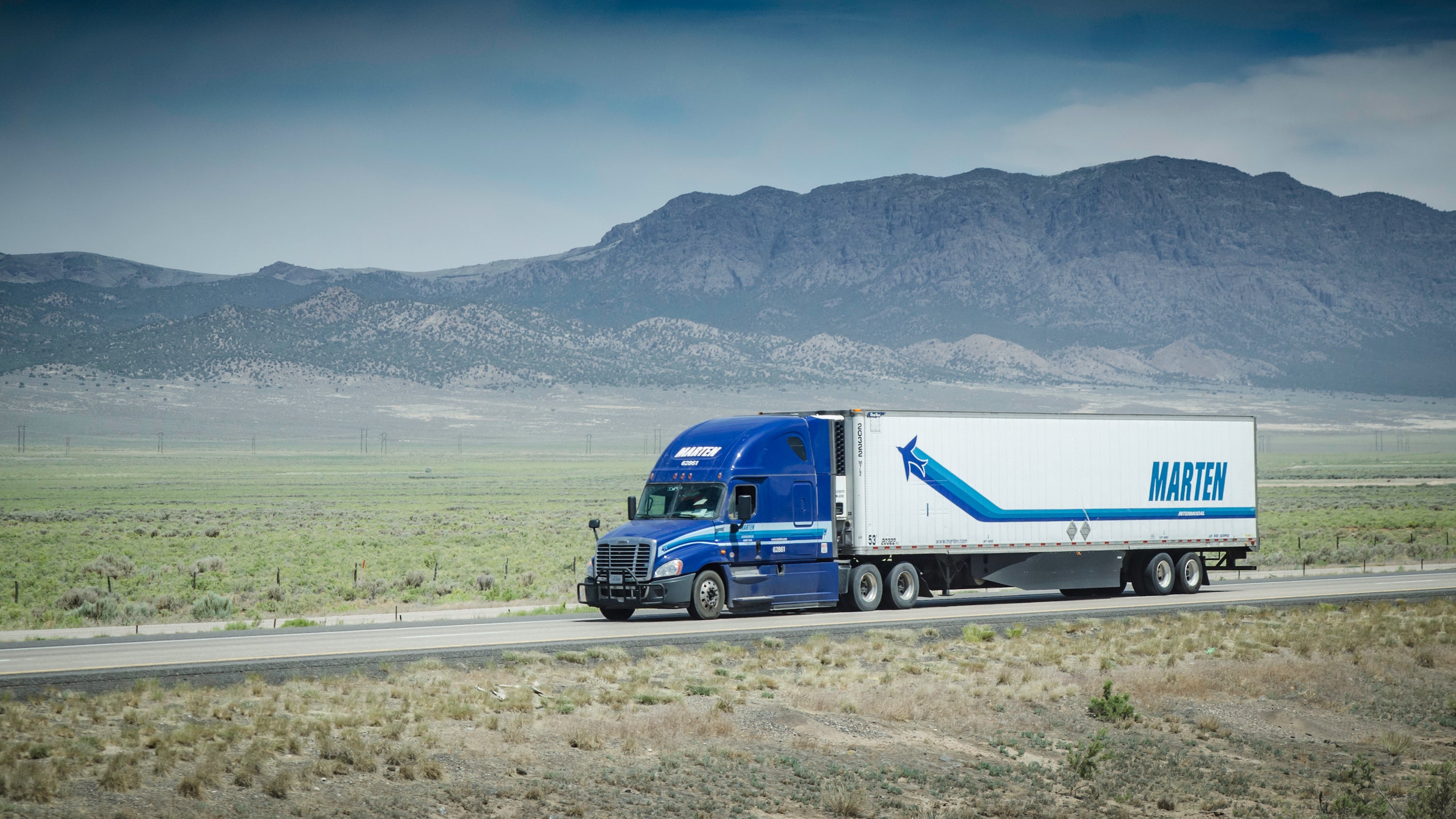 A tractor-trailer of Marten Transport travels on a highway with mountains in the background.