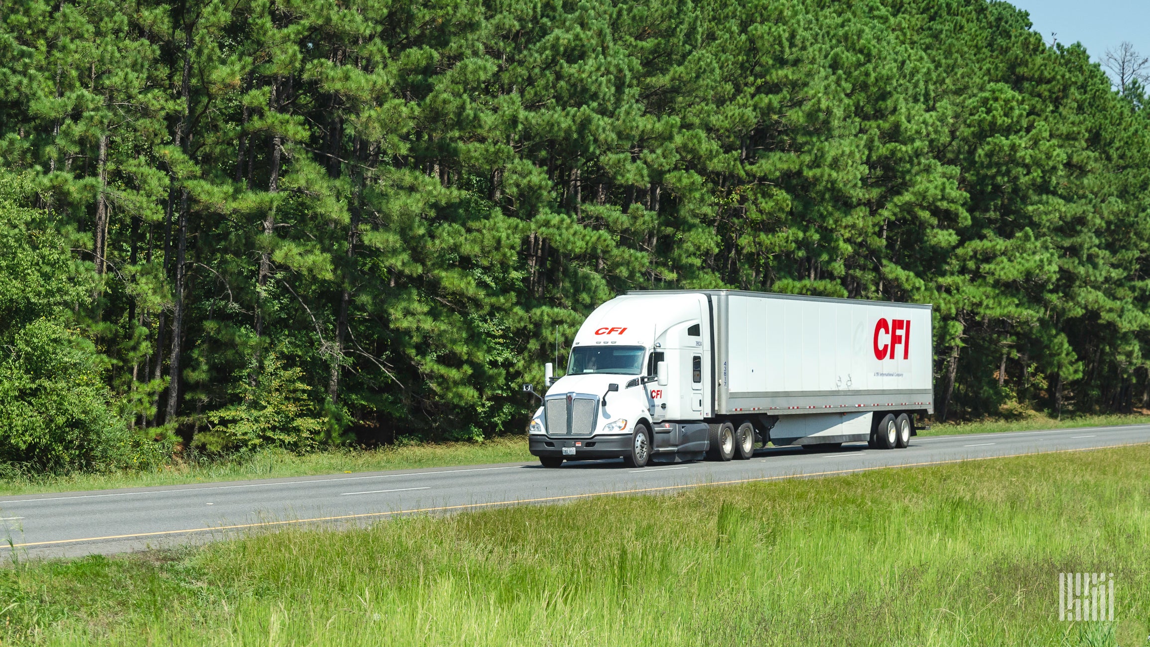 A white tractor-trailer of TFI International carrier CFI traveling on a road with green pine trees in the background.