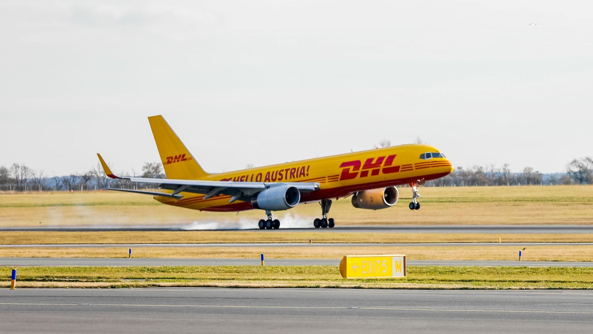 A mustard-colored DHL cargo plane touches down on a runway.