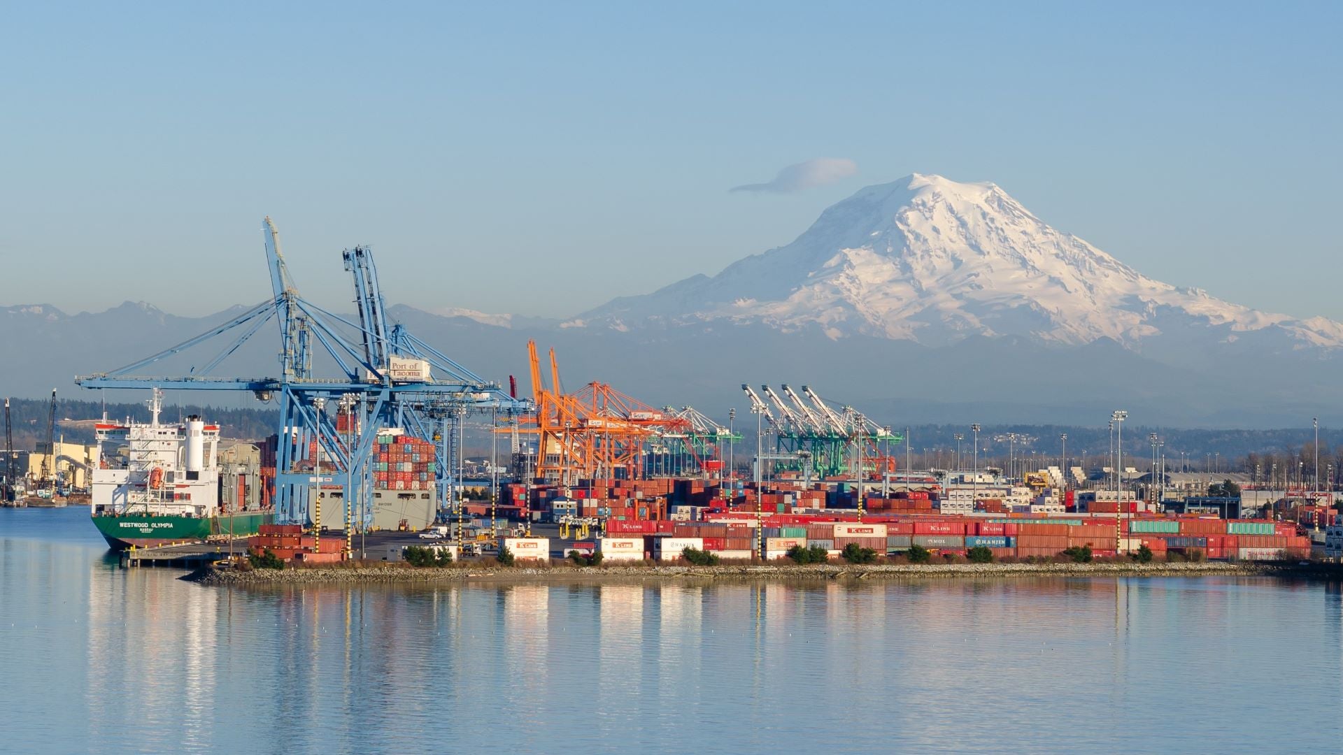 Waterside view of the Port of Tacoma, with a ship at berth, tall cranes and a snow-capped mountain in the background.