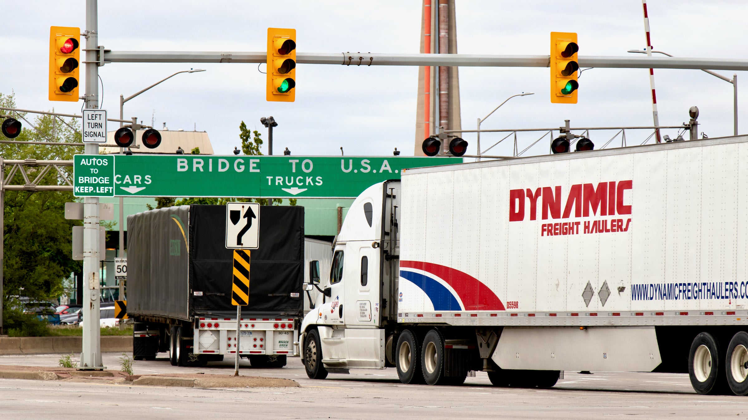 Transport trucks pass under a "Bridge To USA" at the entrance to the Ambassador Bridge, US-Canada border crossing, illustrating an article about the impacts of a COVID-19 vaccine mandate on Canadian truckers.