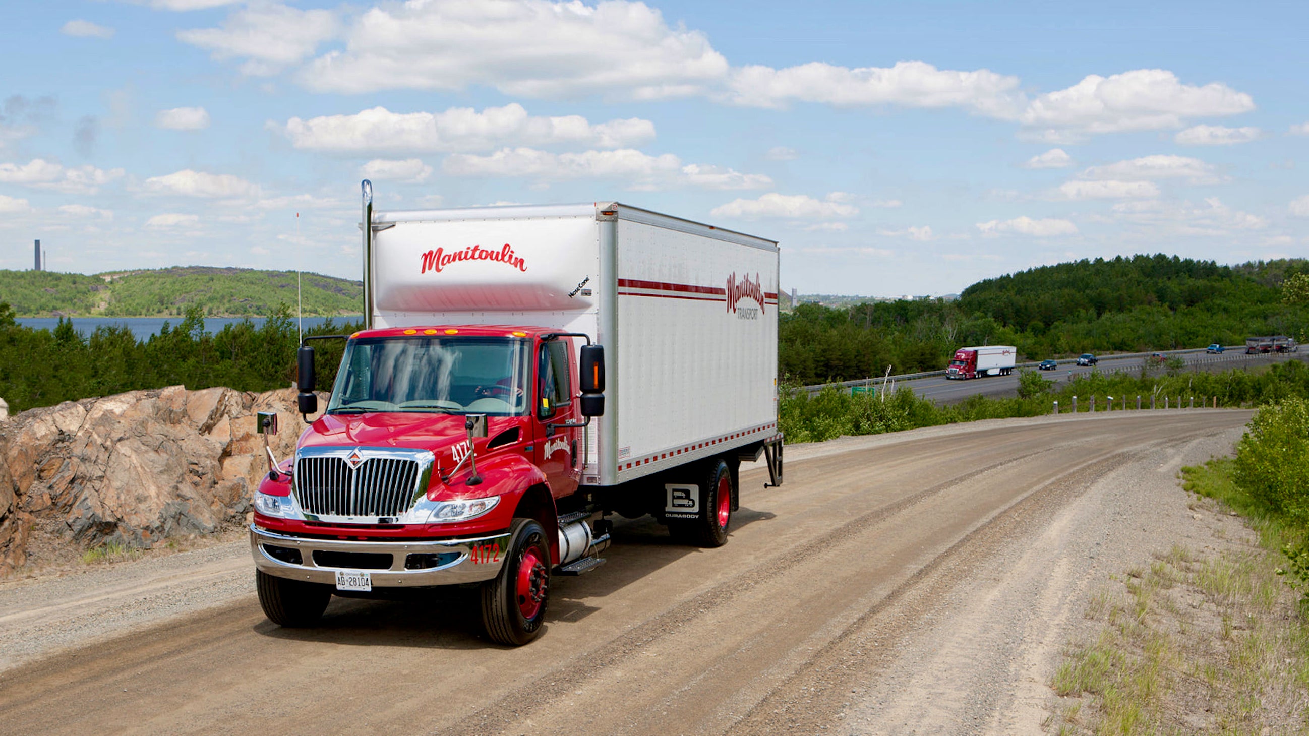 A red and white box truck of Manitoulin Transport on a dirt road with Manitoulin tractor-trailer on a highway the background