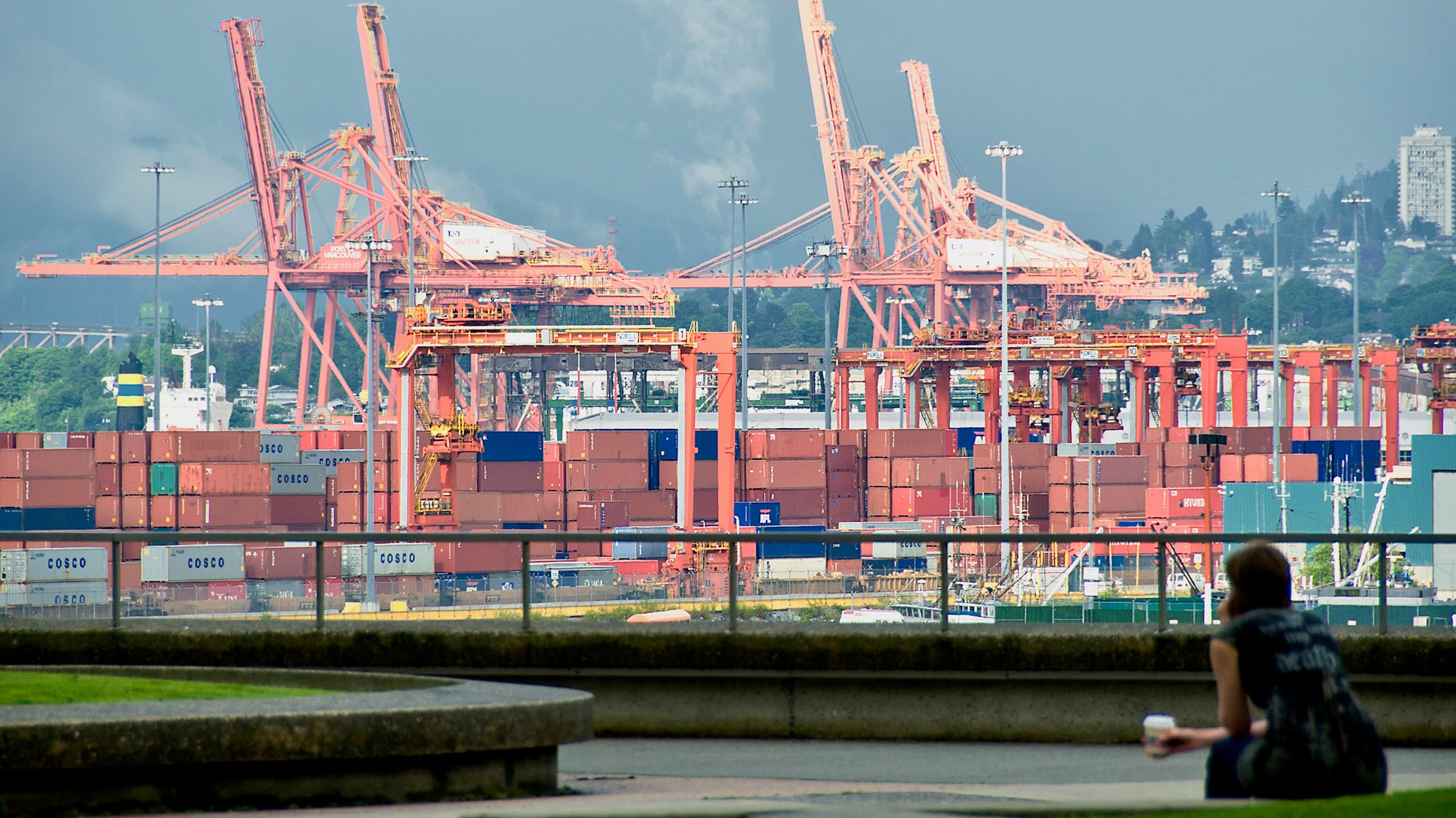 Shipping containers are loaded by cranes at the Port of Vancouver with a woman holding a cup of coffee in the foreground.