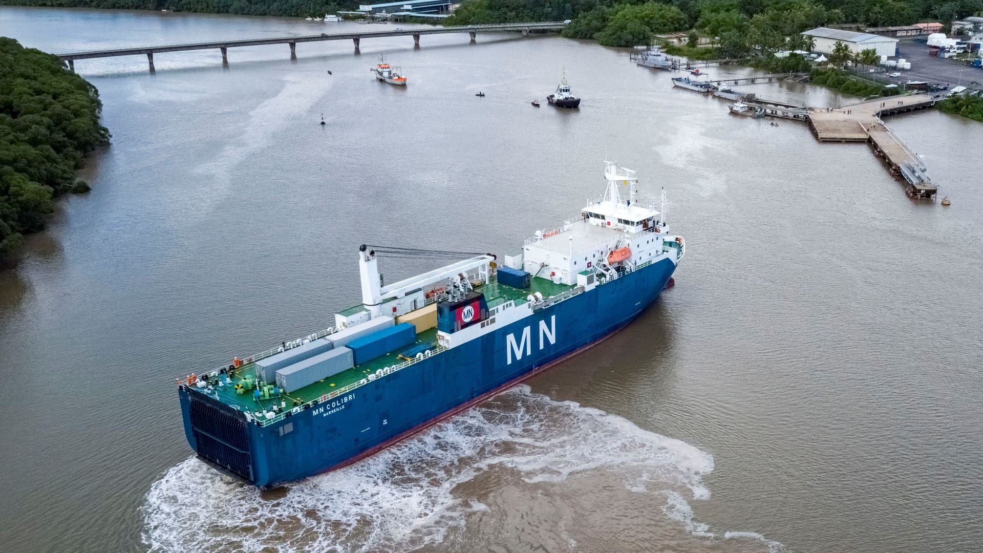 A blue cargo ship prepares to dock at a small river port.