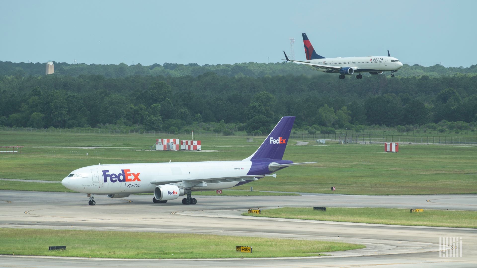 A white and purple FedEx plane on the tarmac as a Delta plane comes in for landing on another runway.