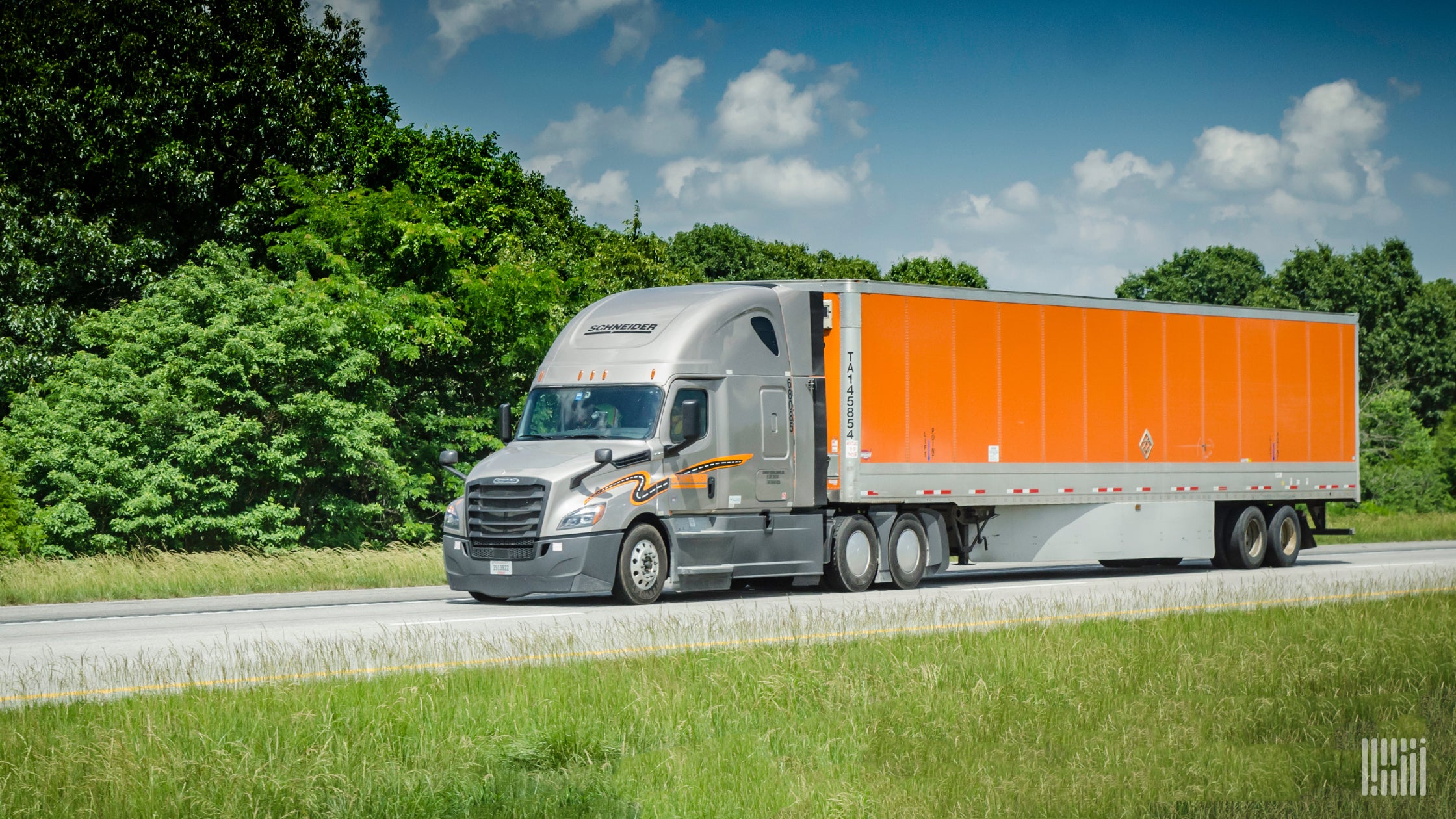 An orange tractor-trailer of Schneider National travels on a road with trees behind it.