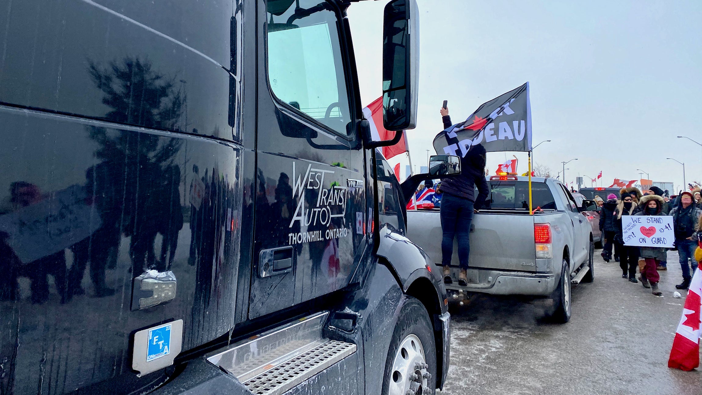 A semi truck moves forward as people holding signs and Canadian flags cheer on.