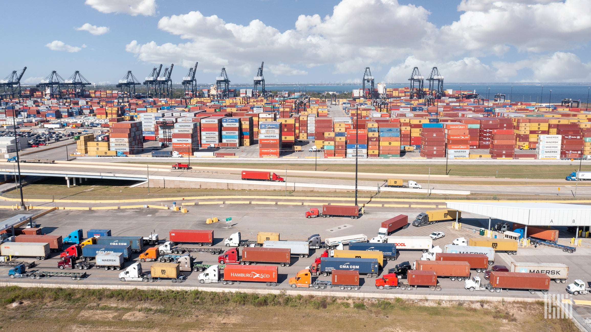 Wide view of a container marine terminal with cranes, containers and trucks on a sunny day.