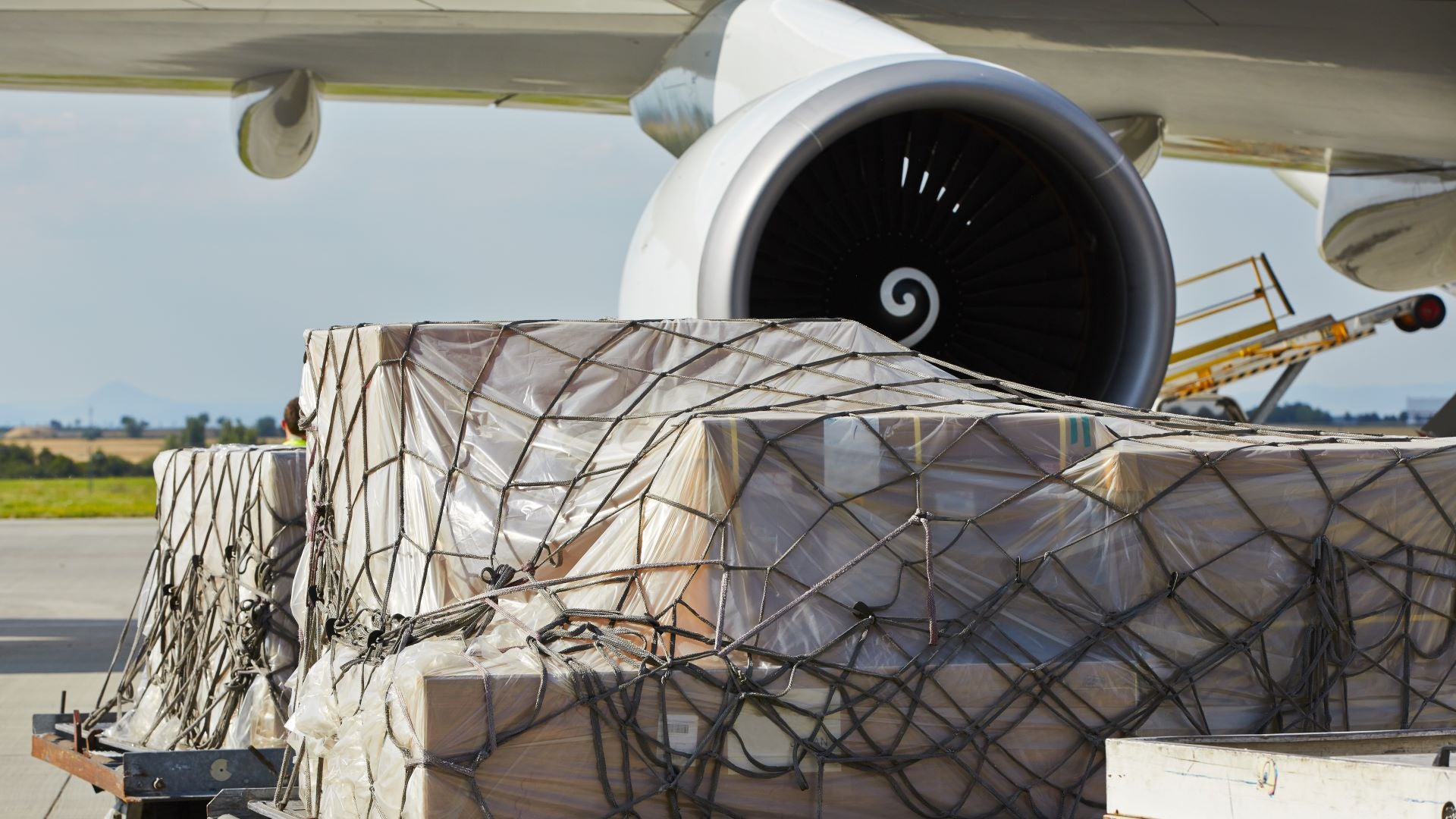 A large cargo pallet with netting on tarmac in front of an aircraft's engine.