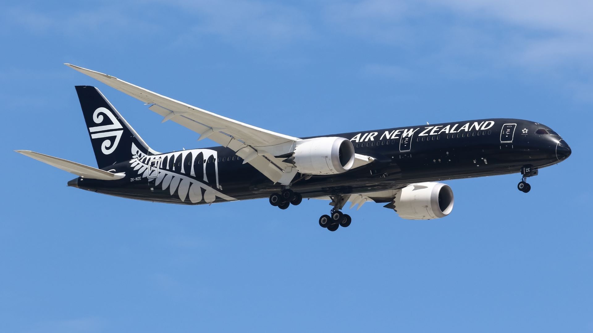 An all-black Air New Zealand plane with wheels down on approach in blue sky.