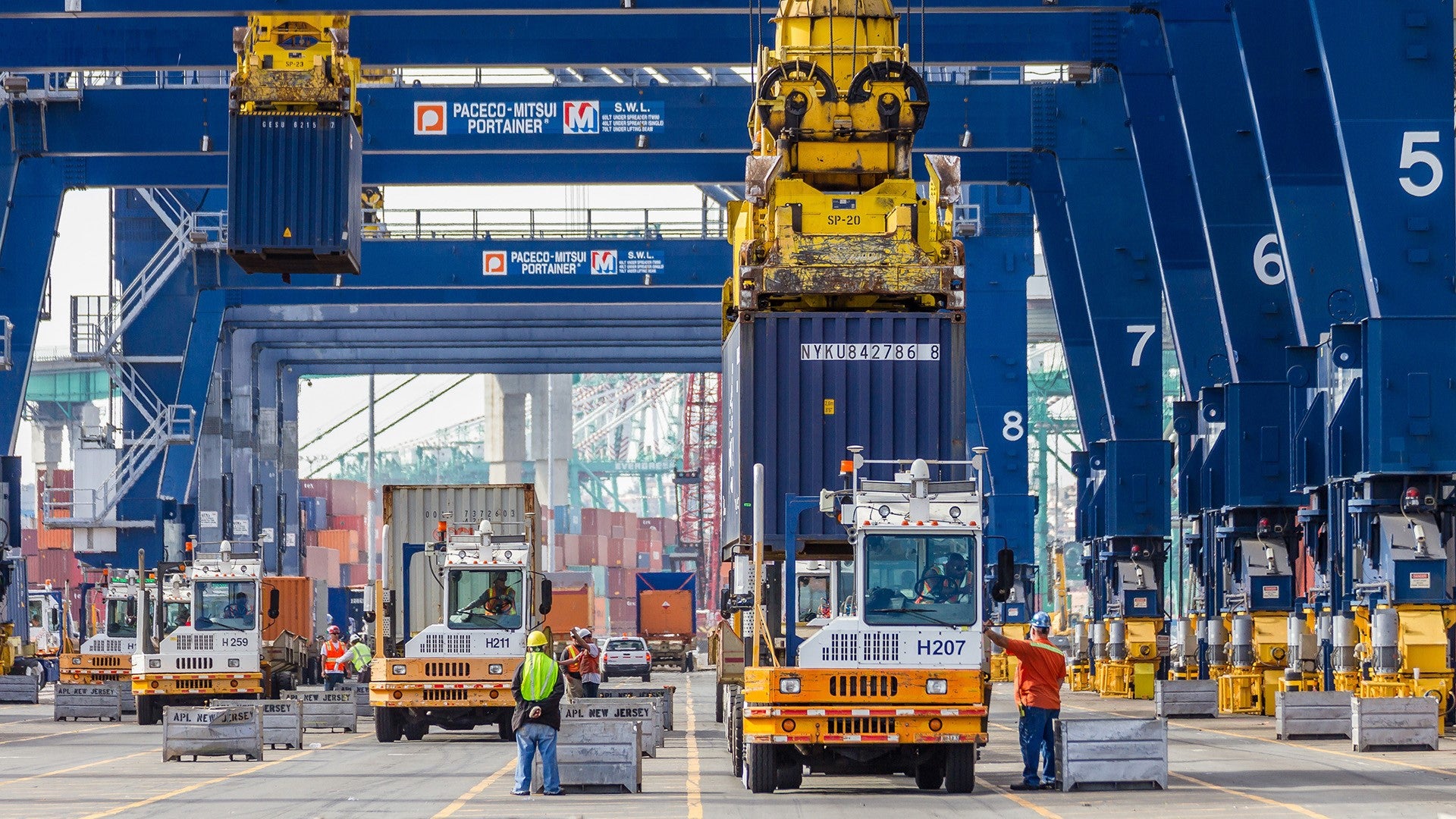 Workers and trucks under giant ship cranes at a port.