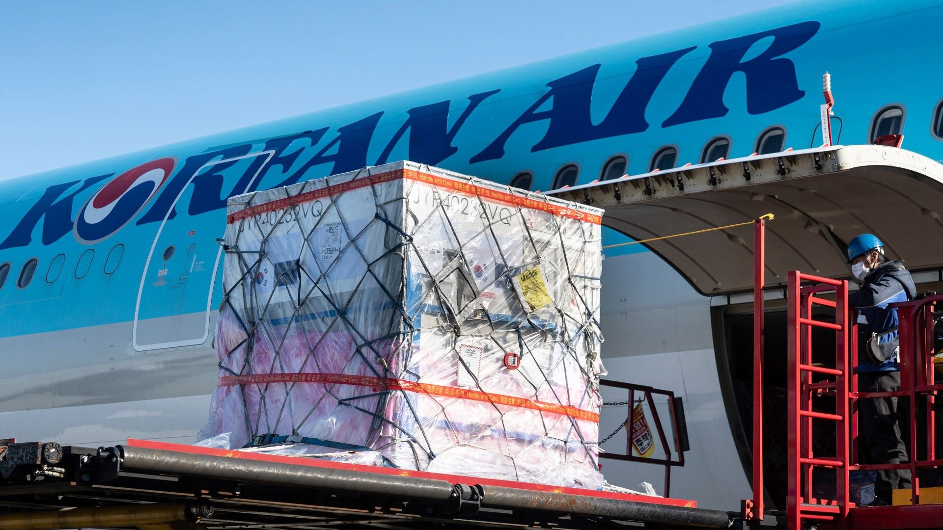 A large silver container on a lift goes in the side of a pale blue Korean Air jet.