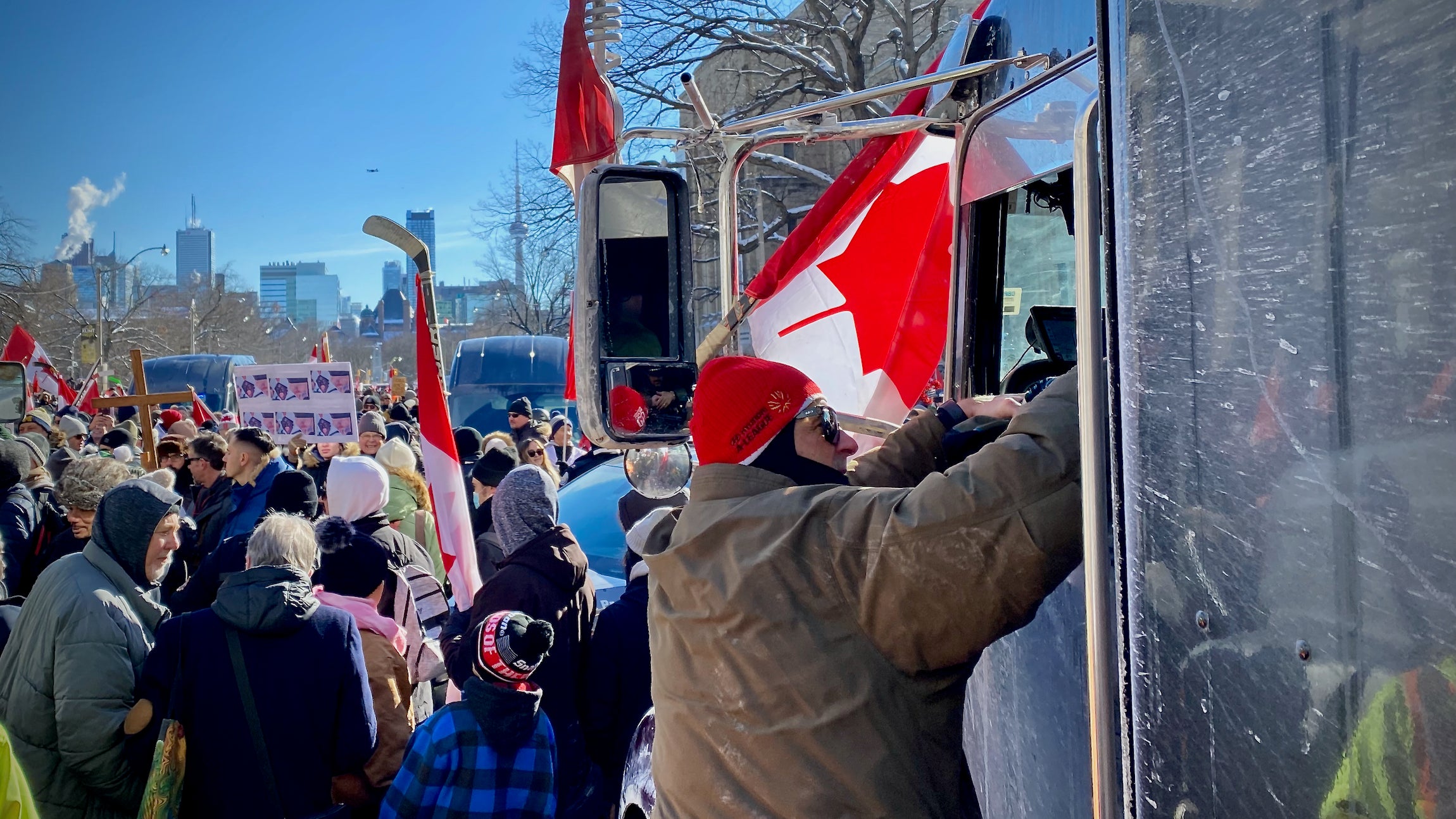 A man talks through the window of a truck while holding a Canadian flag as other protesters crowd around him.