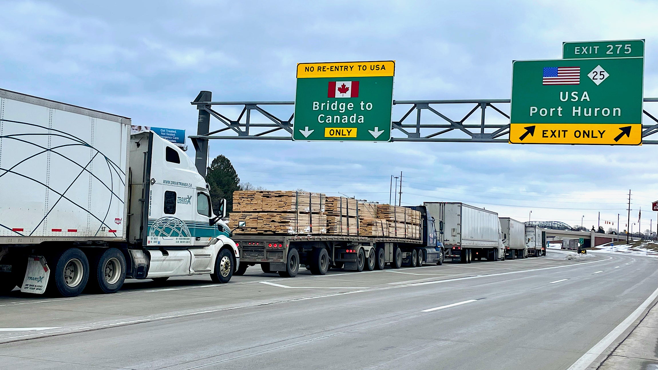 A line of trucks move on a road where a sign reads bridge to Canada