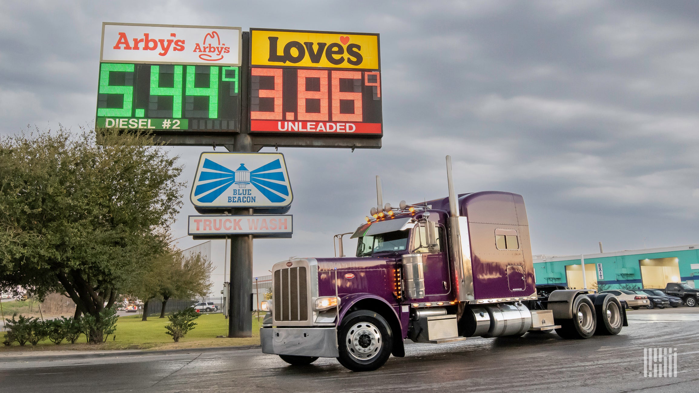 A red semi-truck is parked in front of a sign displaying the prices of diesel and regular gas