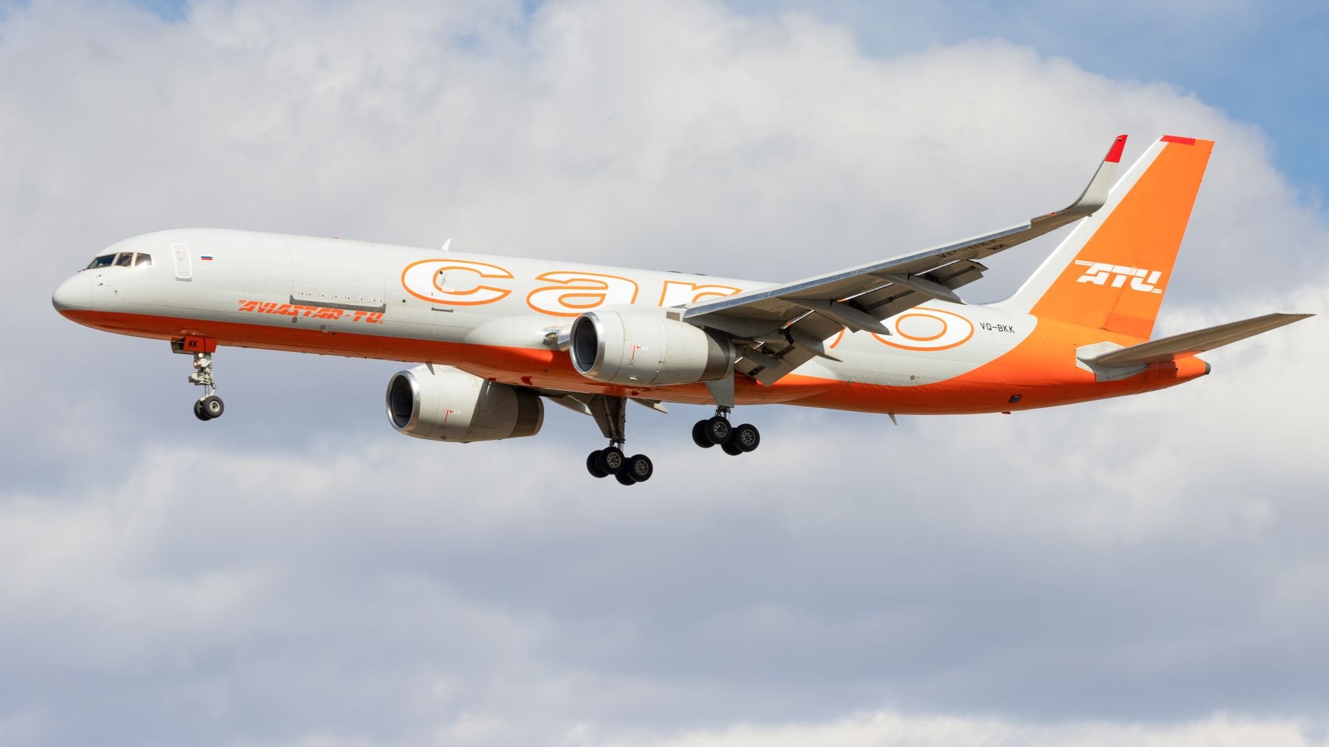 A gray Aviastar cargo plane with an orange tail on approach to an airport with wheels down.