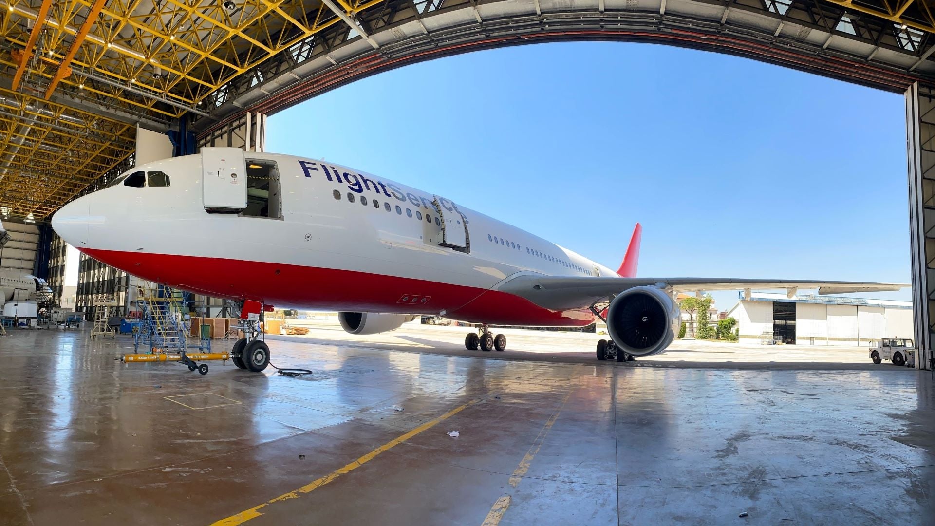 A white commercial jet inching into a large storage hanger with blue sky in the background. The plane belongs to AELF FlightService.