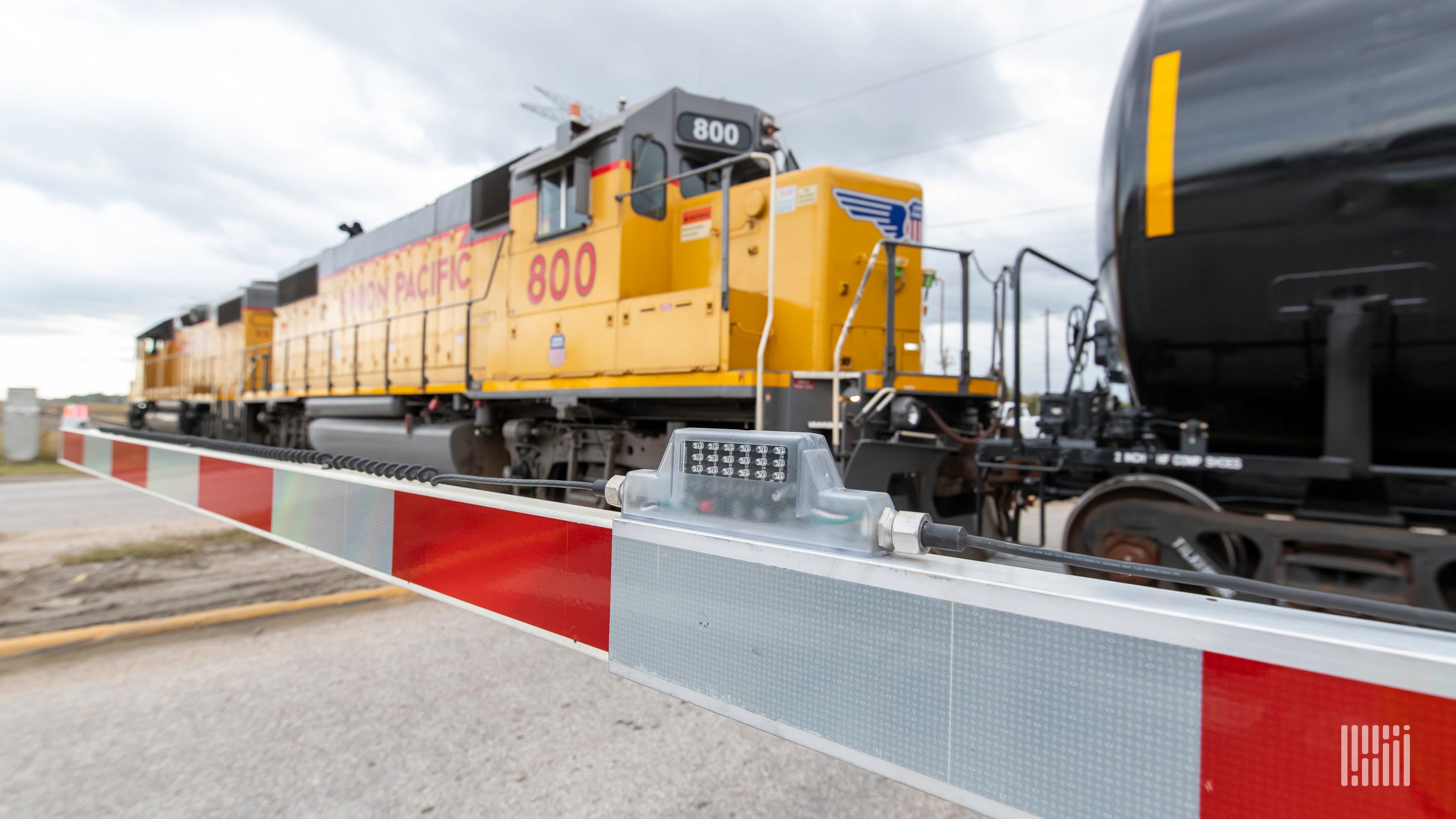 A photograph of a freight train traveling by a rail crossing. A guard rail on the road is down to protect motorists.