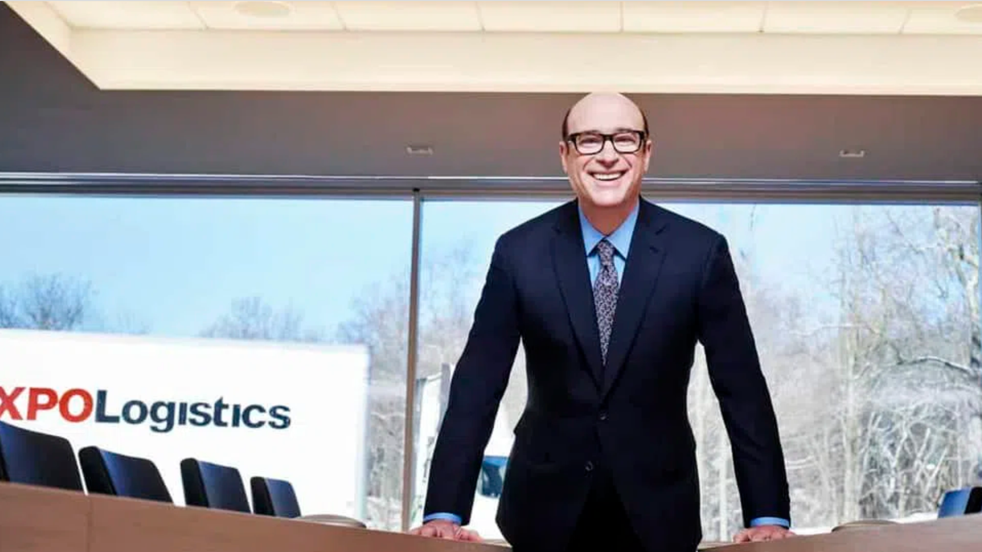 A man with glasses and wearing a suit stands behind a table with a the logo of XPO Logistics visible