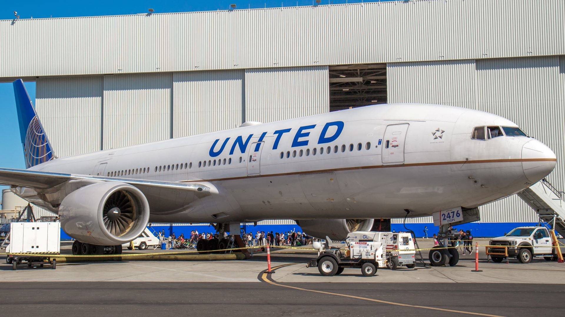 A white United Airlines 777 plane in front of a maintenance hanger with carts around it. The 777s have been out of service for more than a year to resolve a safety issue with the engines.