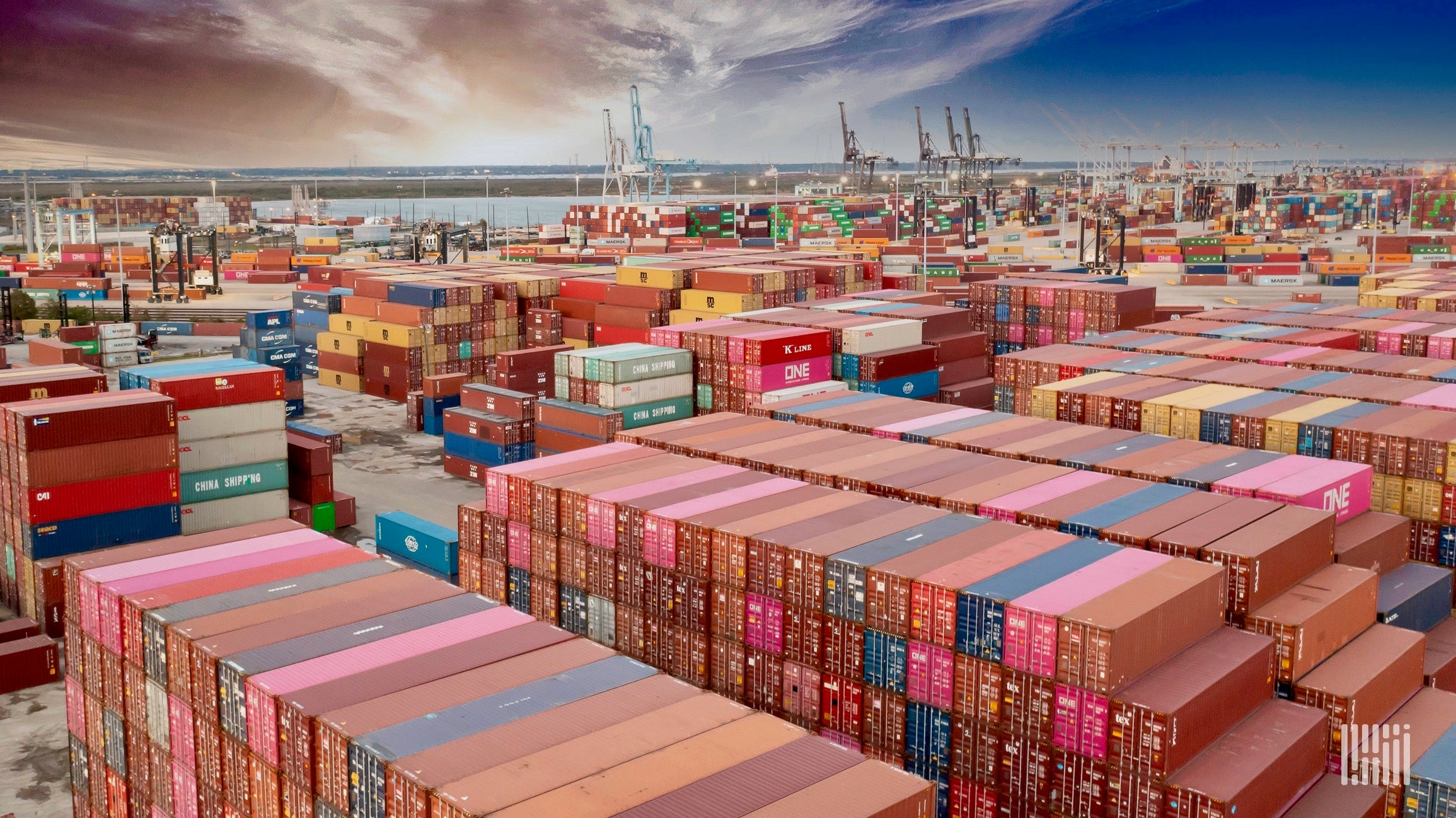 Thousands of intermodal containers of various colors are stacked at a port while storm clouds are in the background.