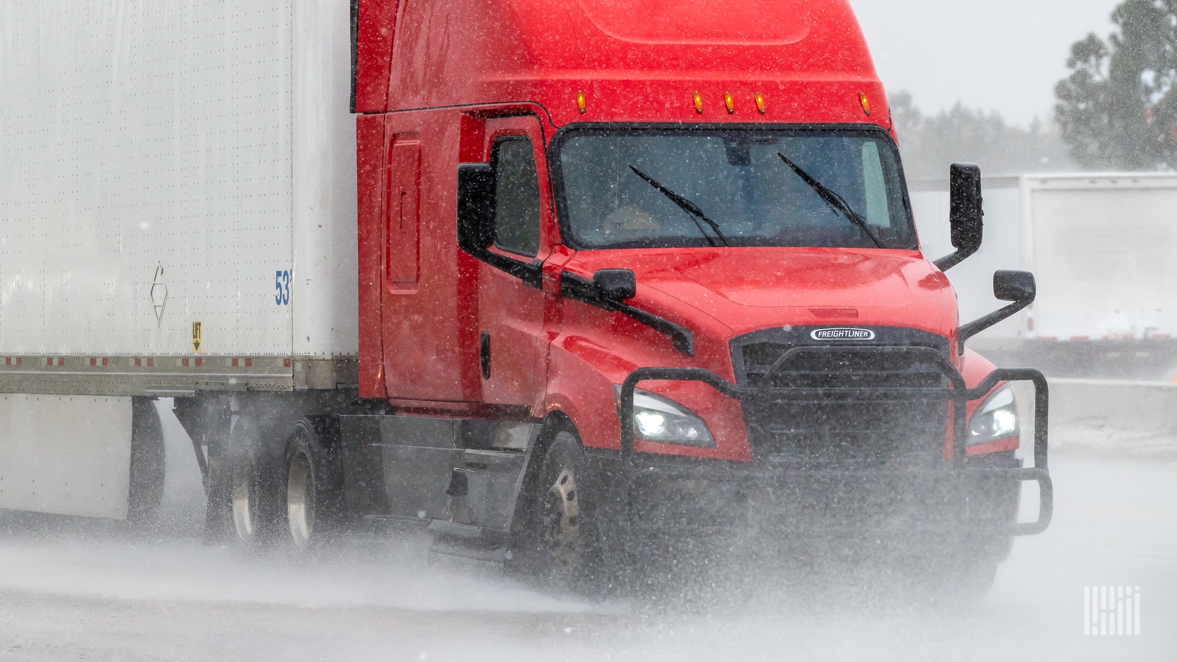 A red semi truck with a white trailer drives through rain