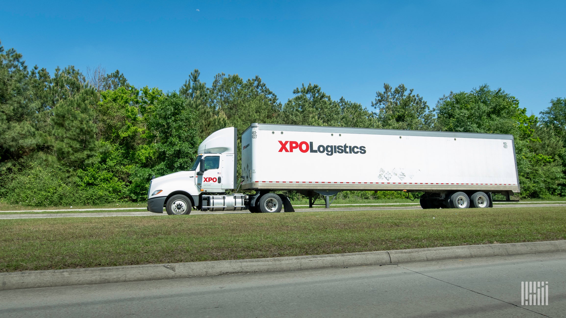 A white XPO Logistics tractor-trailer travels on a major road with a green landscape and blue skies in the background.