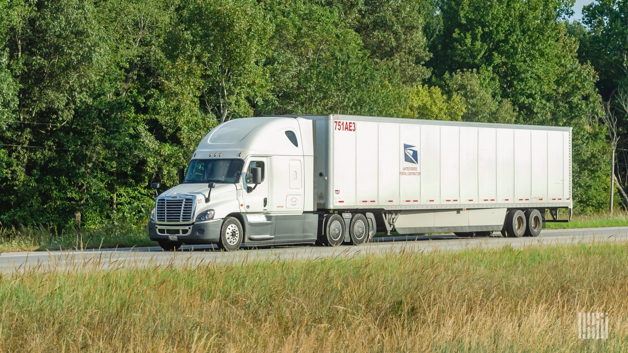 A tractor-trailer with the logo of the US Postal Service traveling on a road