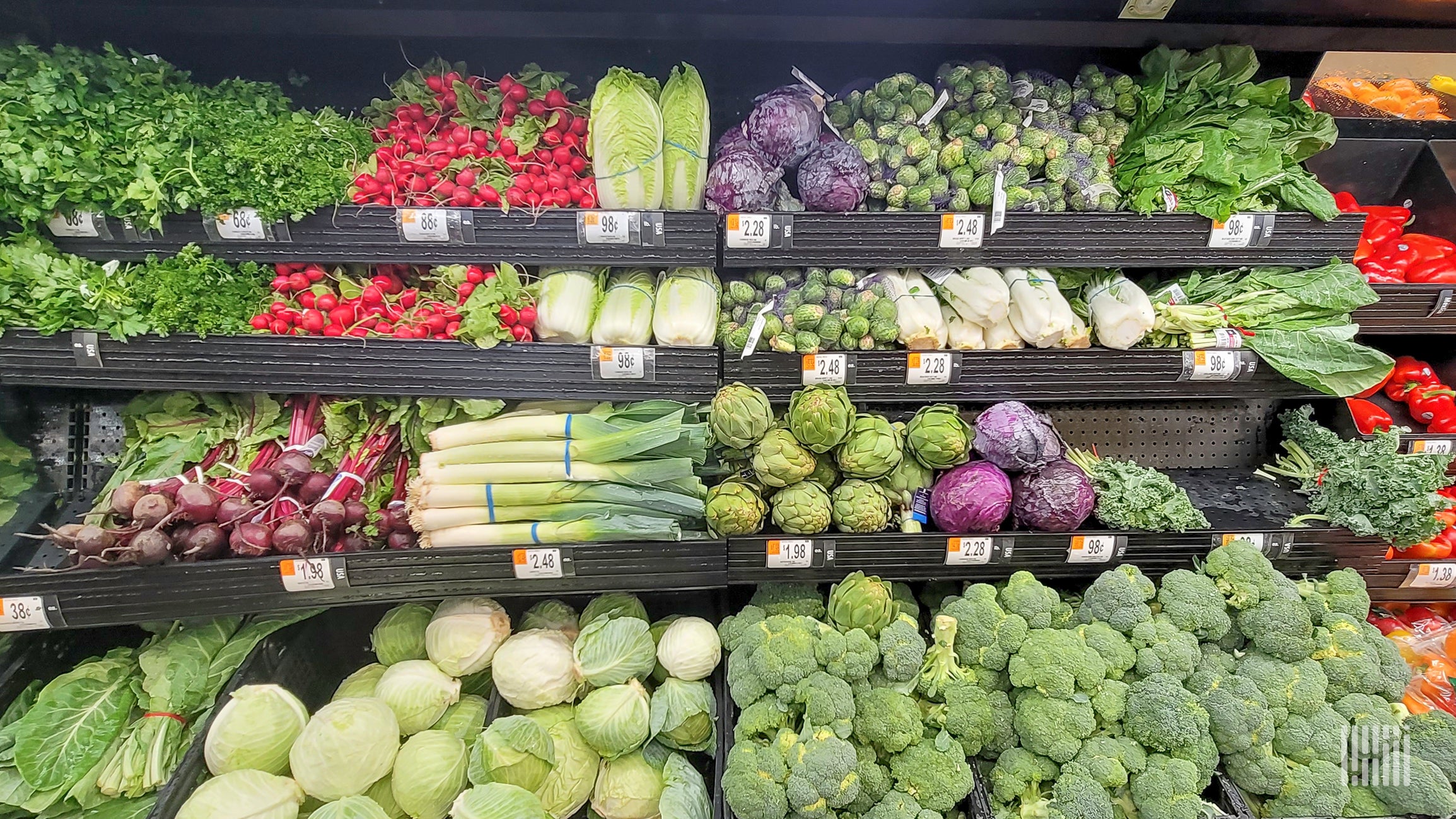 A produce section of a grocery store shows foods such as broccoli, celery, cabbage, radishes, and lettuce.