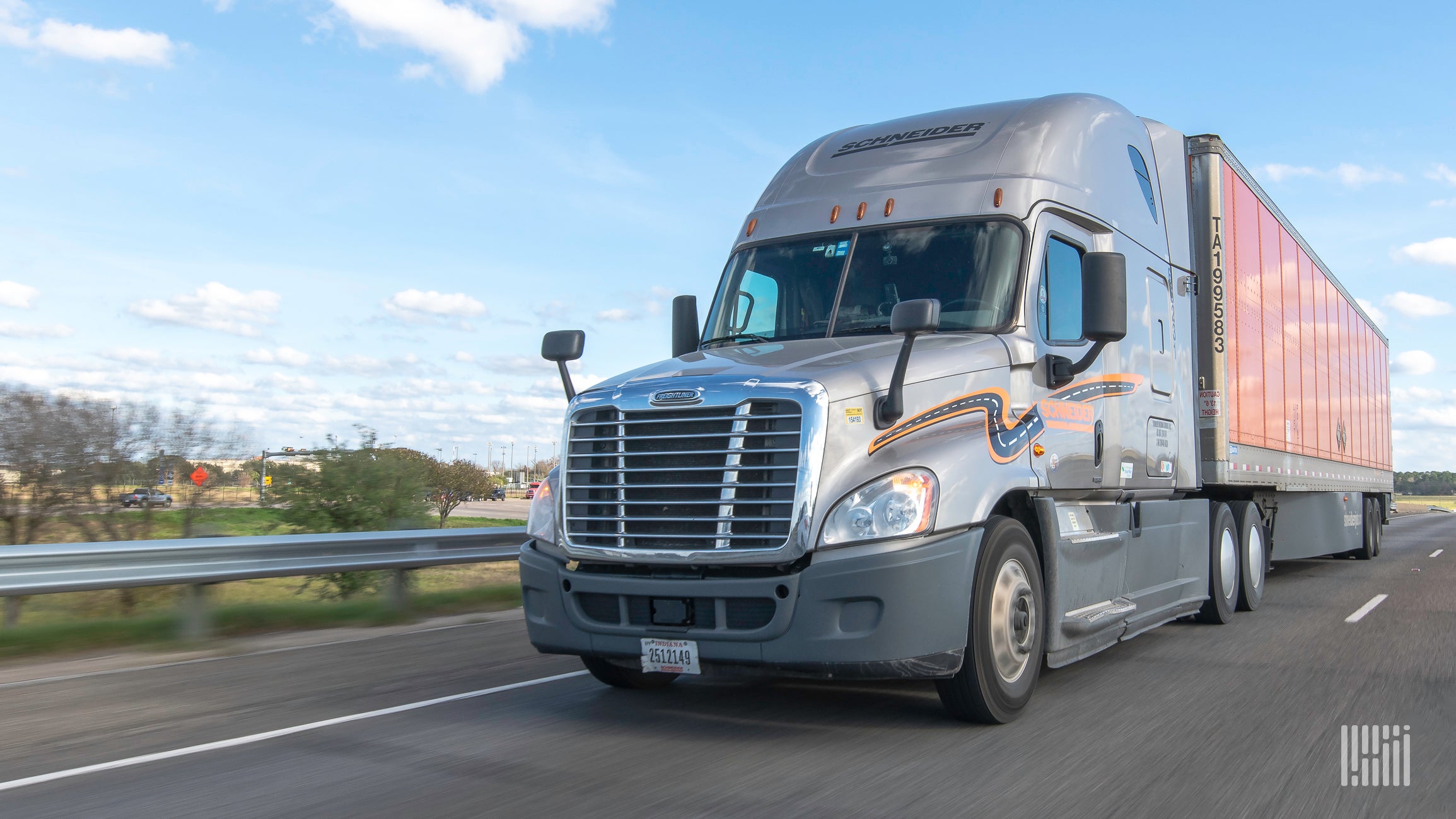 A grey Schneider tractor trailer is traveling on a road with trees and a blue sky behind.