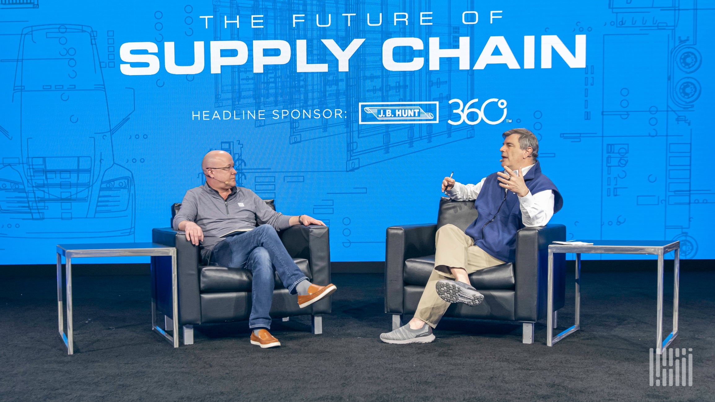 Two men sitting in chairs on a stage and talking to each other.