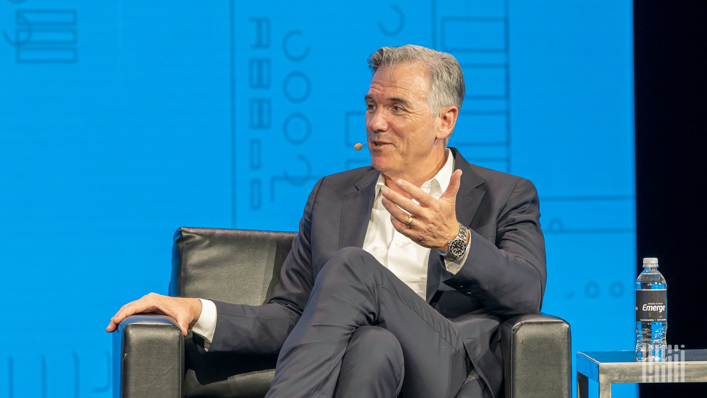 A man sitting on a leather chair talks to another person with one arm extended.