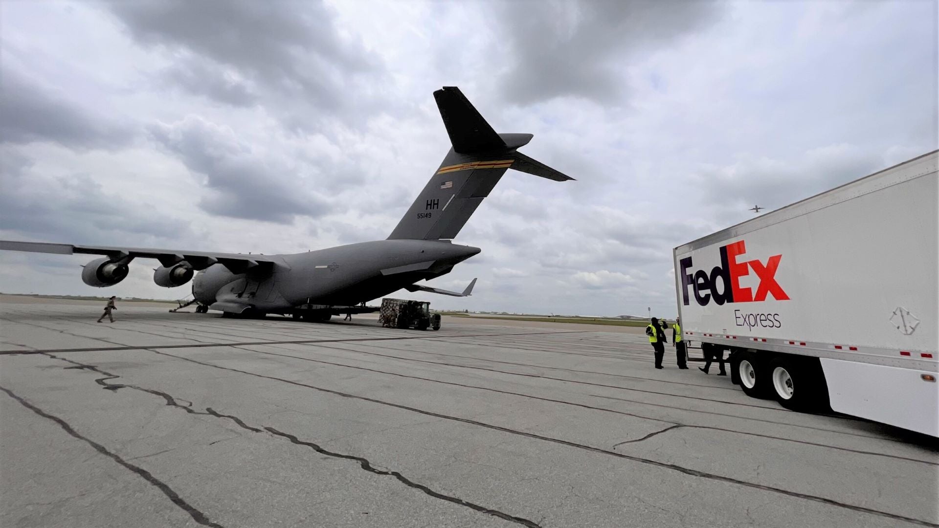 A white FedEx truck on the tarmac behind a large military cargo jet on a cloudy day.