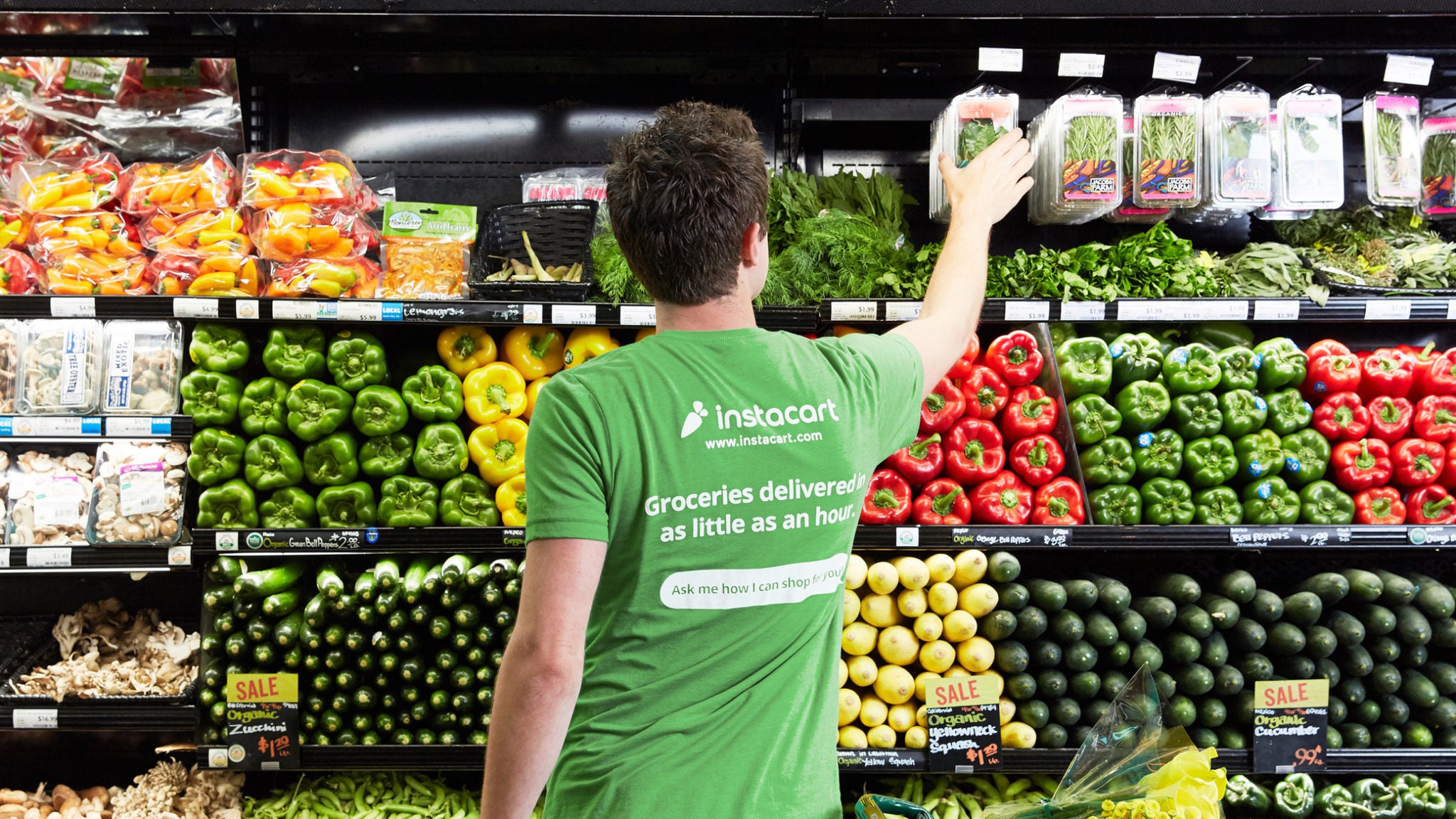 Woman wearing Instacart shirt reaches for vegetables in a grocery store aisle.