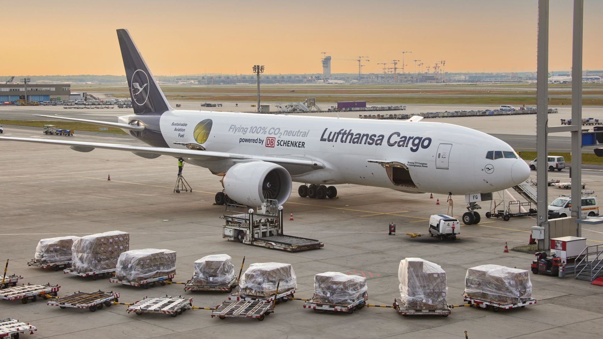 A large Lufthansa cargo jet surrounded by shipping containers on the tarmac with an early dawn sunshine in the background.