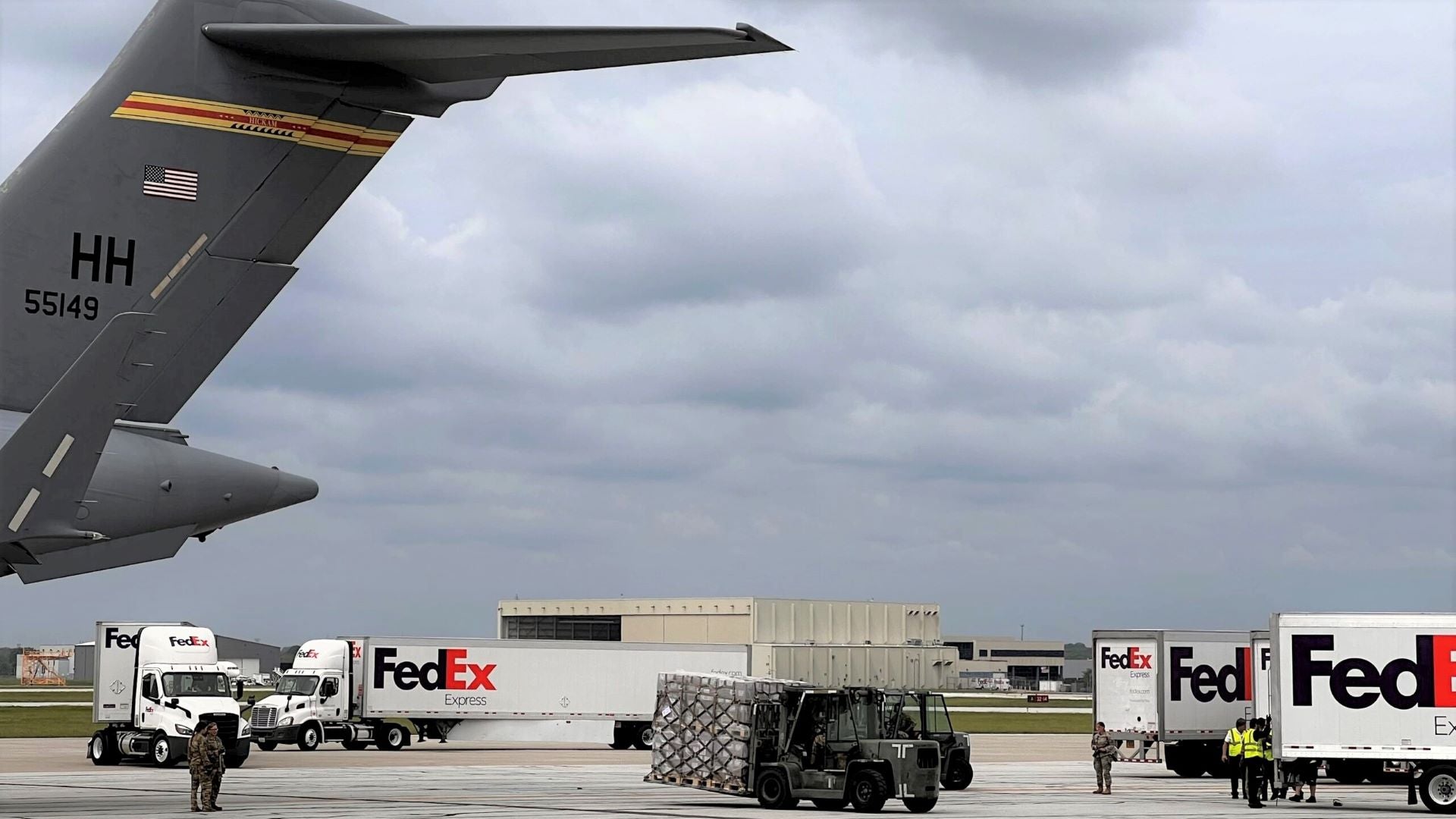 A tail of a military plane and FedEx trucks shown on an airport tarmac as a tractor moves pallets.