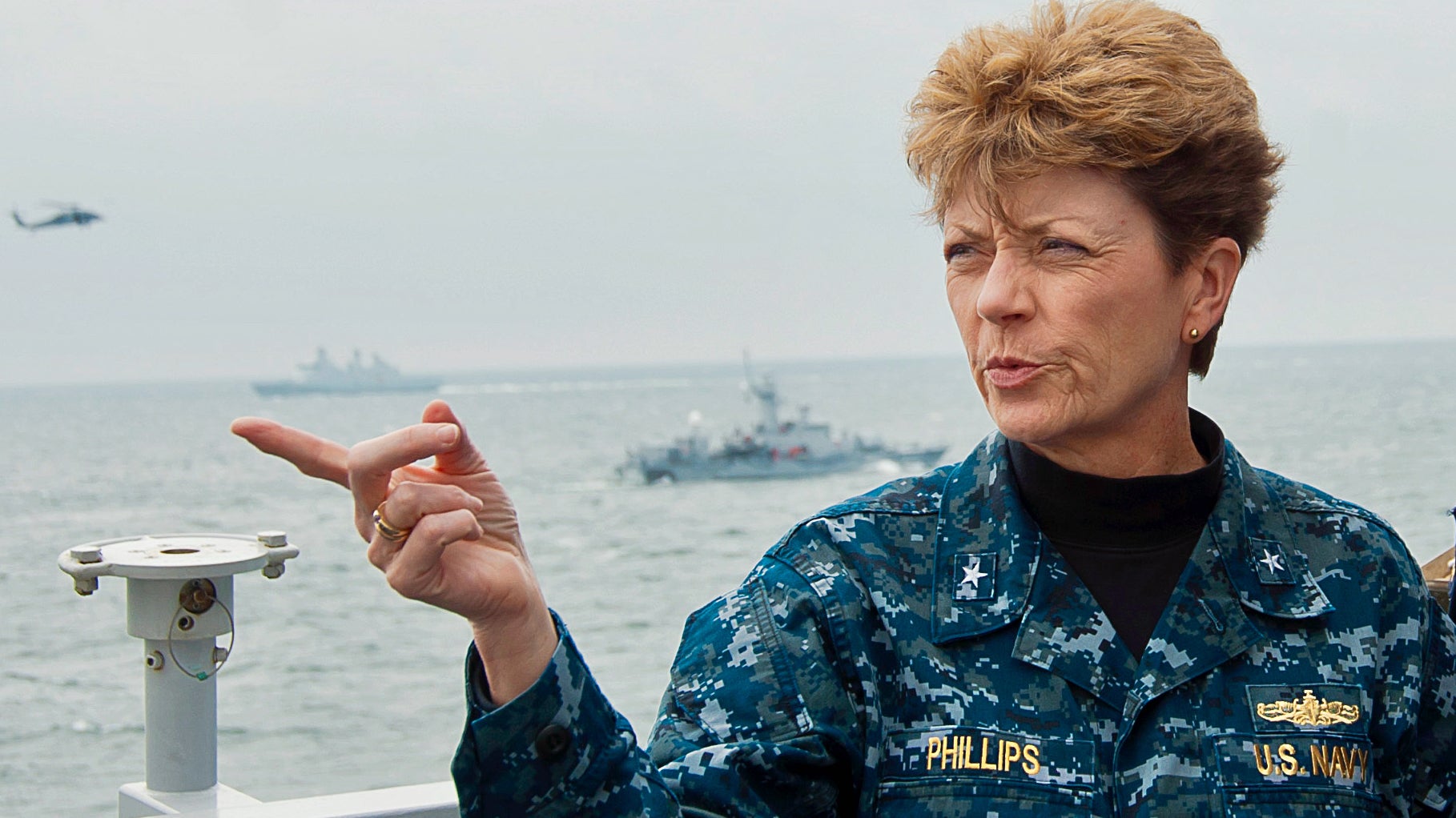 A woman in a blue Navy camouflage uniform with the name Phillips sewn on it, points while on the deck of a ship,