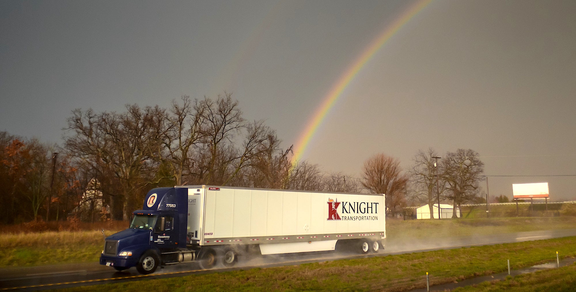 A tractor-trailer with the logo of Knight Transportation drives on a road with storm clouds and a rainbow behind it, illustrating a downturn in the trucking industry.