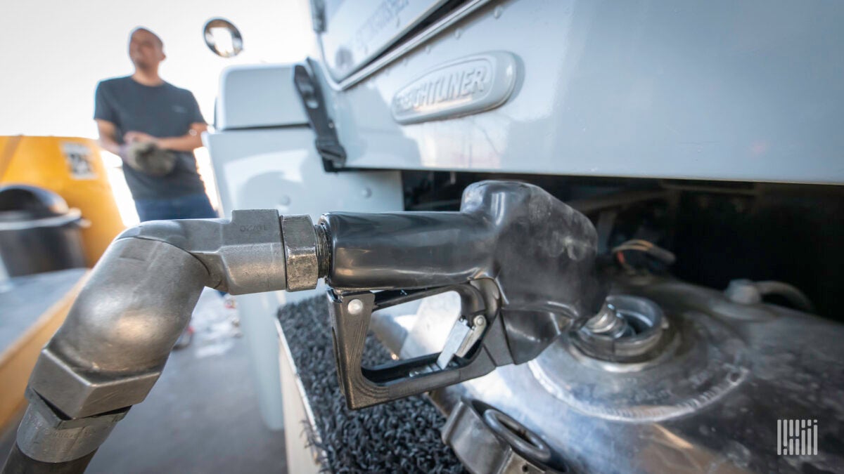 The nozzle of a diesel fuel pump is inserted into the tank of a commercial truck as its driver looks on the bankground.