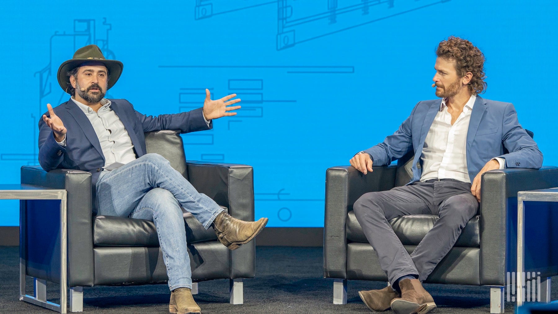 Two men sitting in leather chairs talking, as the one on the left gestures with his arms out.