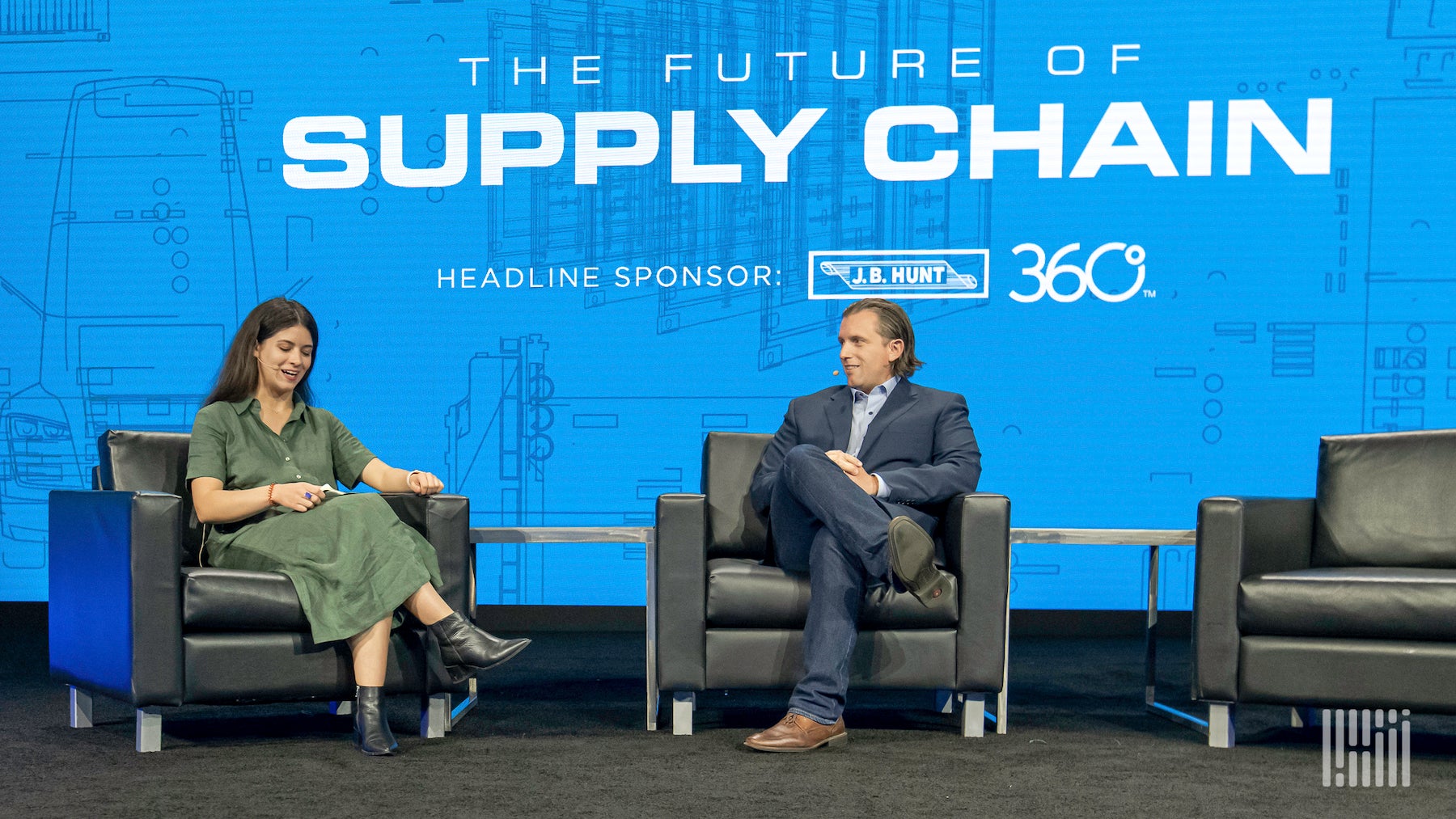 A man and woman sitting in leather chairs with a blue screen behind them with the words The Future of the Supply Chain written on it.