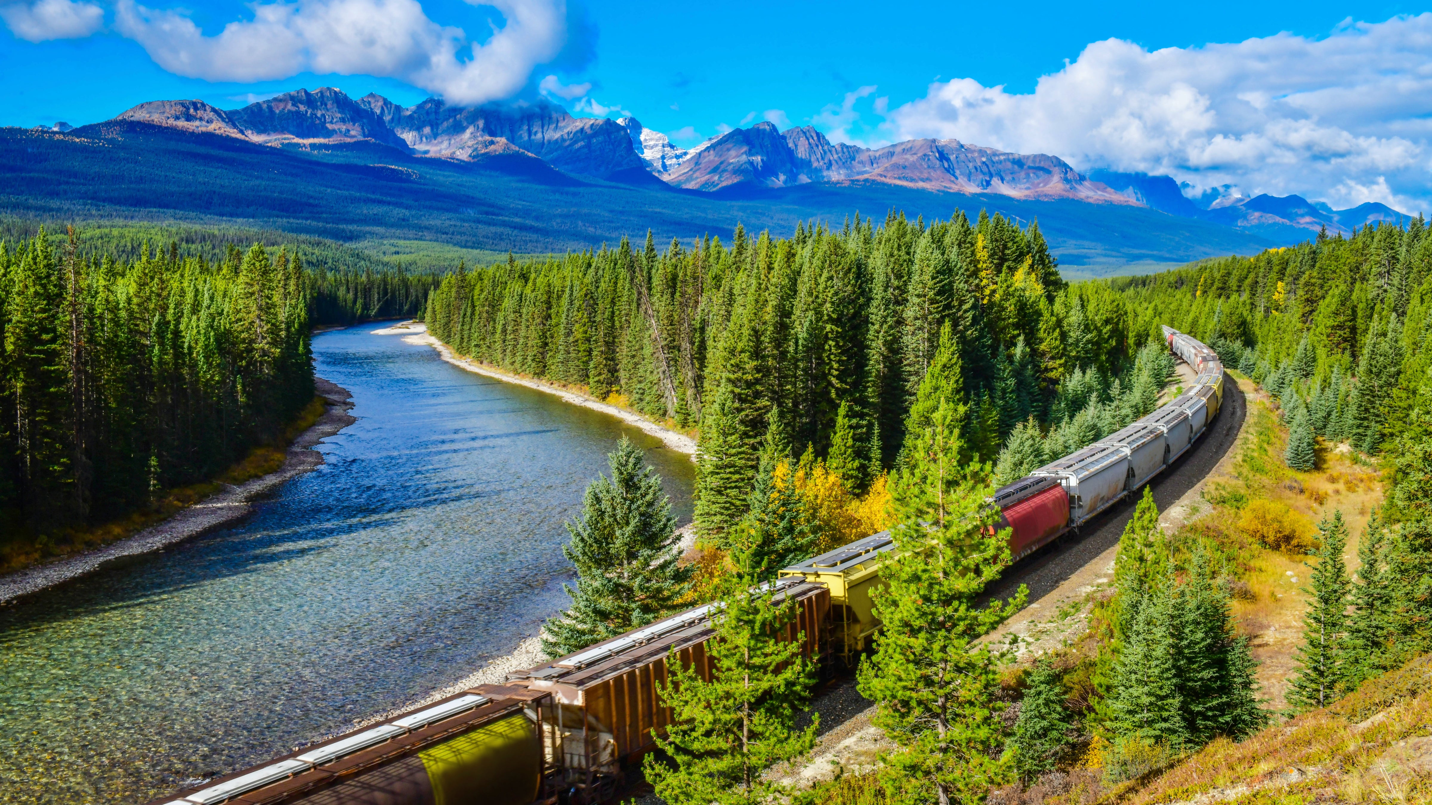 A grain train weaves through a Canadian evergreen forest. A snow-capped mountain range is in the background.