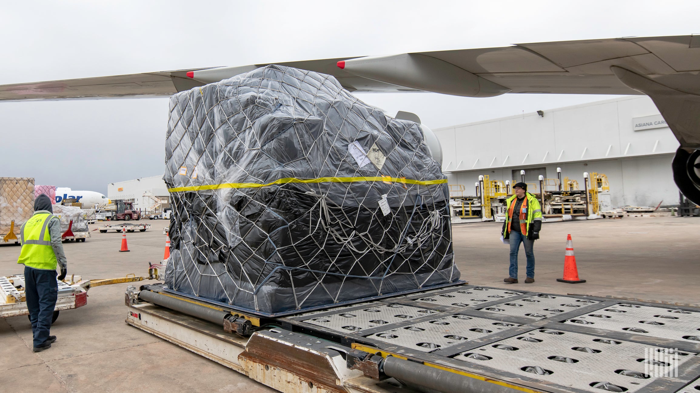 Workers wearing yellow vests maneuver a large freight pallet under the wing of a jetliner.