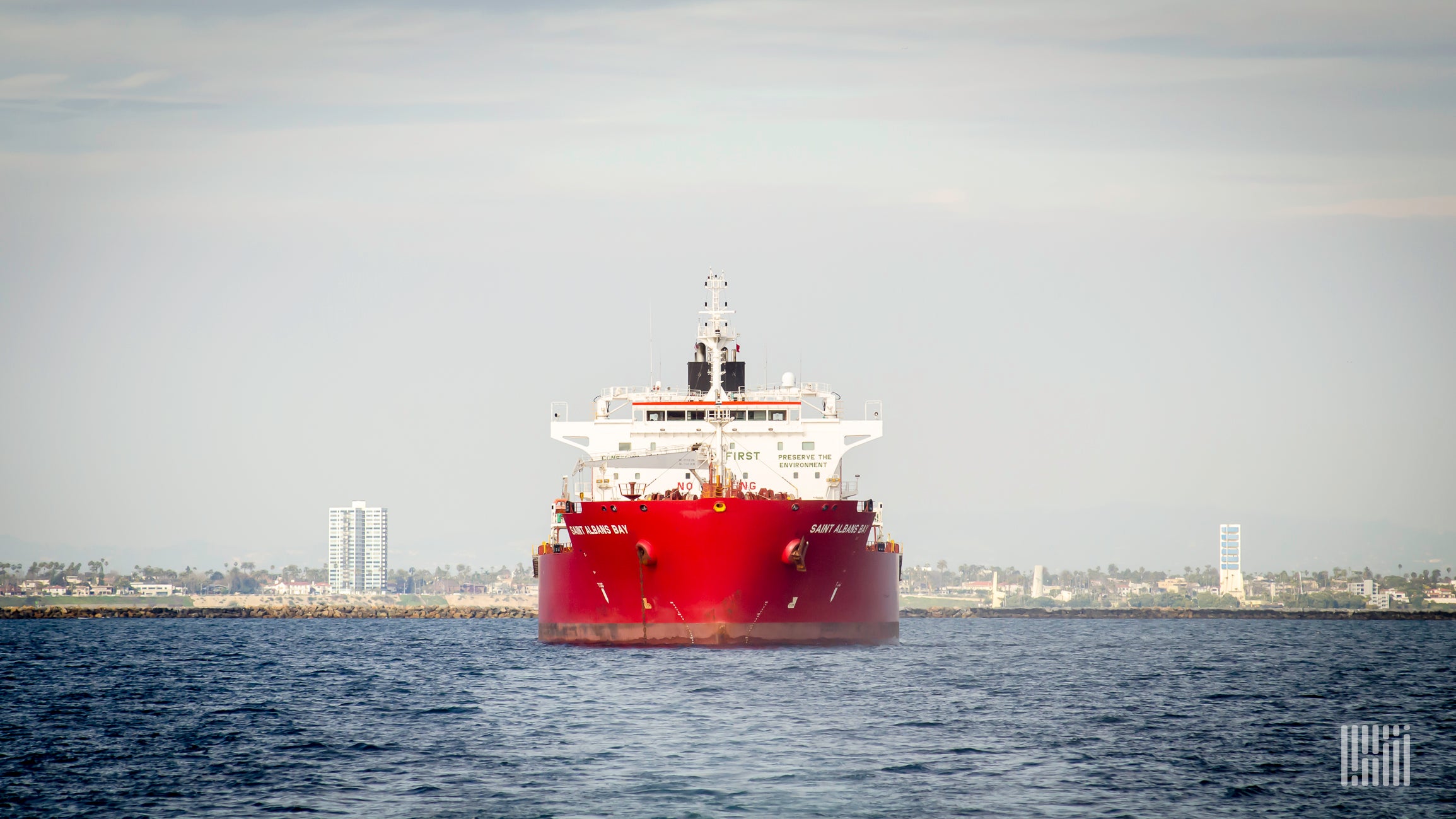 A red and white container ship is traveling on the ocean with the shoreline in the background.