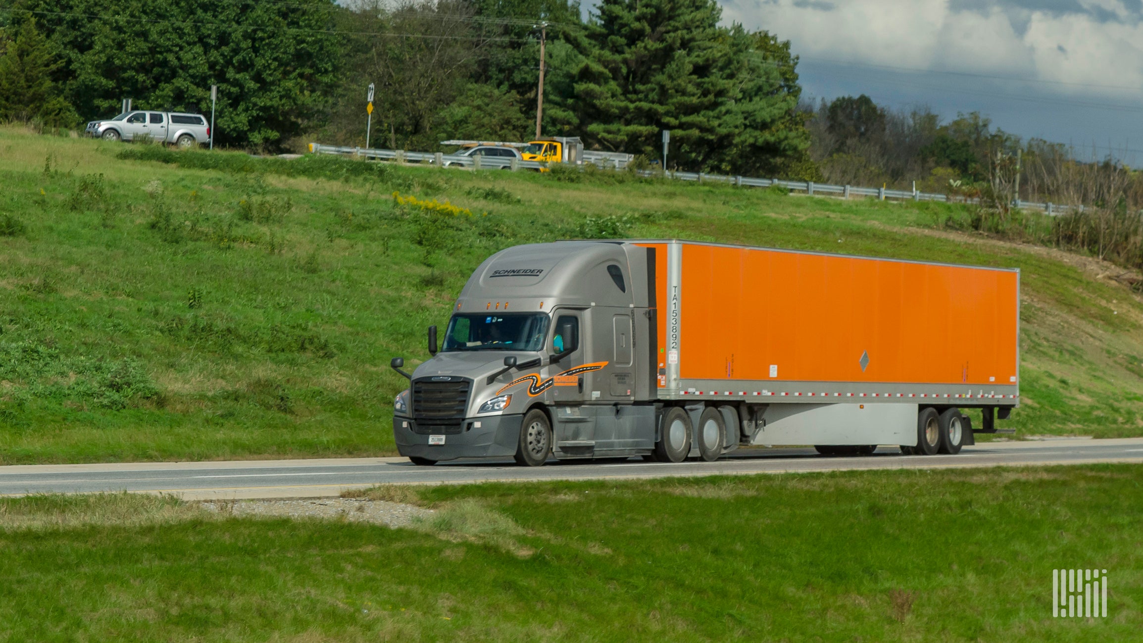 A grey and orange Schneider truck travels down a road green trees and grass in the background.