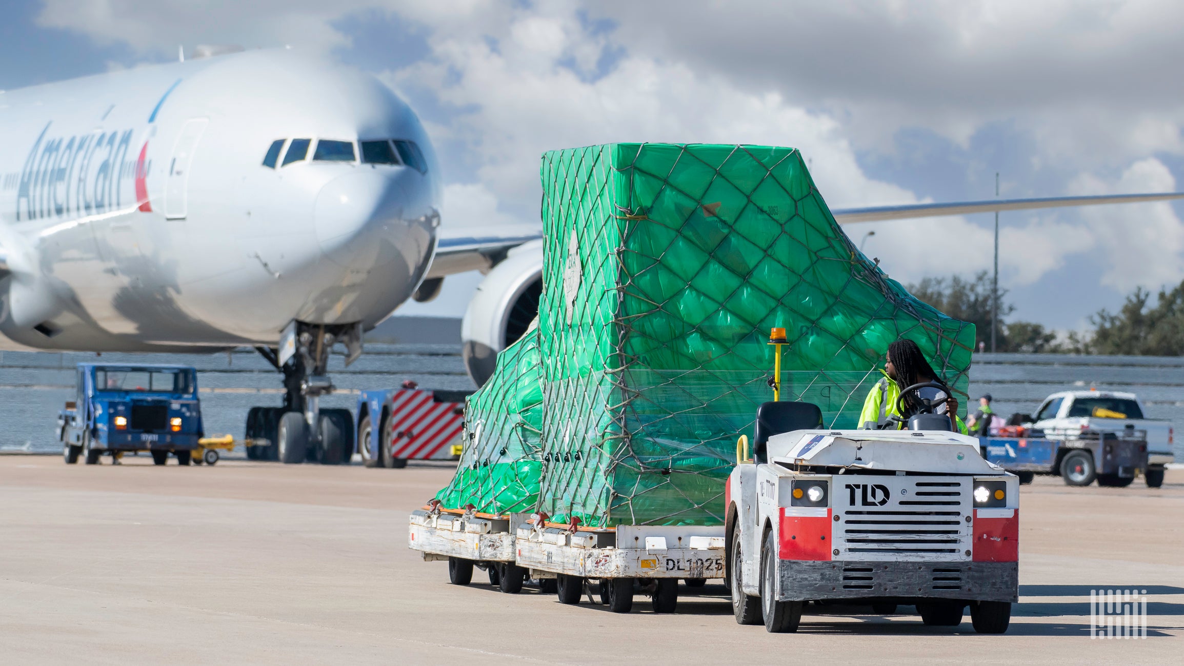 An airport tug pulls a large cargo pallet in front of an American Airlines jet.