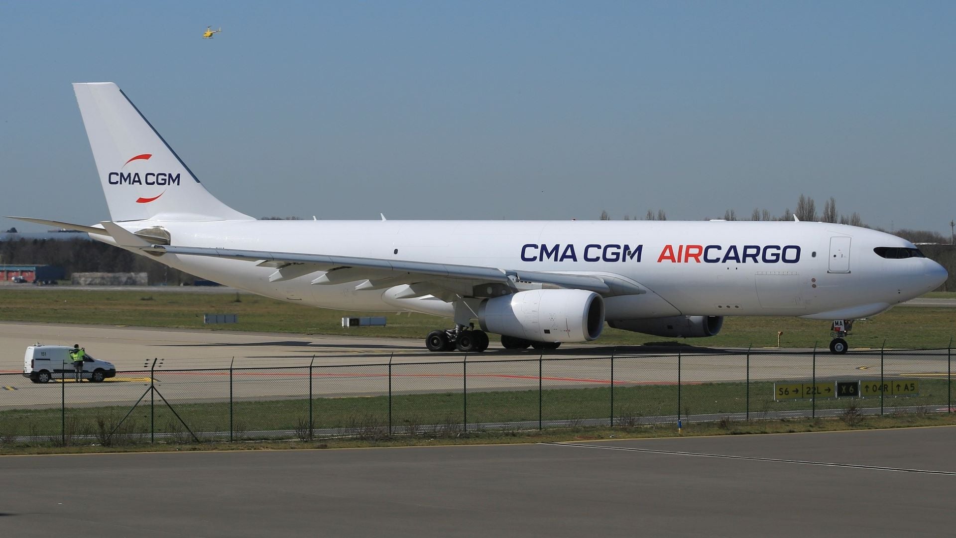 An all-white jet with CMA CGM Air Cargo lettering near the perimeter fence of an airport.