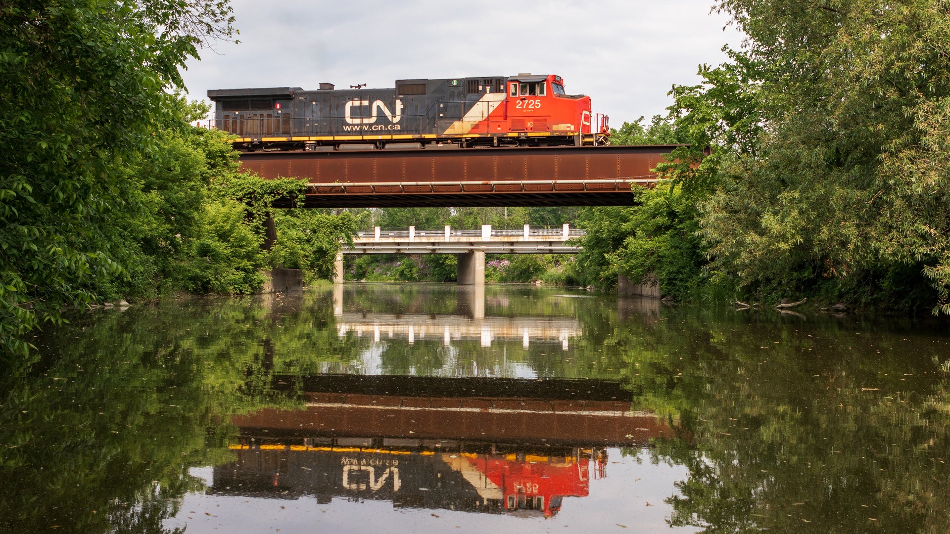 A CN train travels on a bridge and the reflection of the CN locomotive is in the lake underneath the bridge.