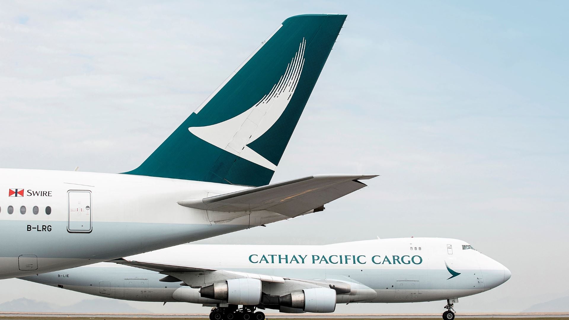 Tail end of a Cathay Pacific jet with green tail, with a Cathay 747 cargo jet in the background.