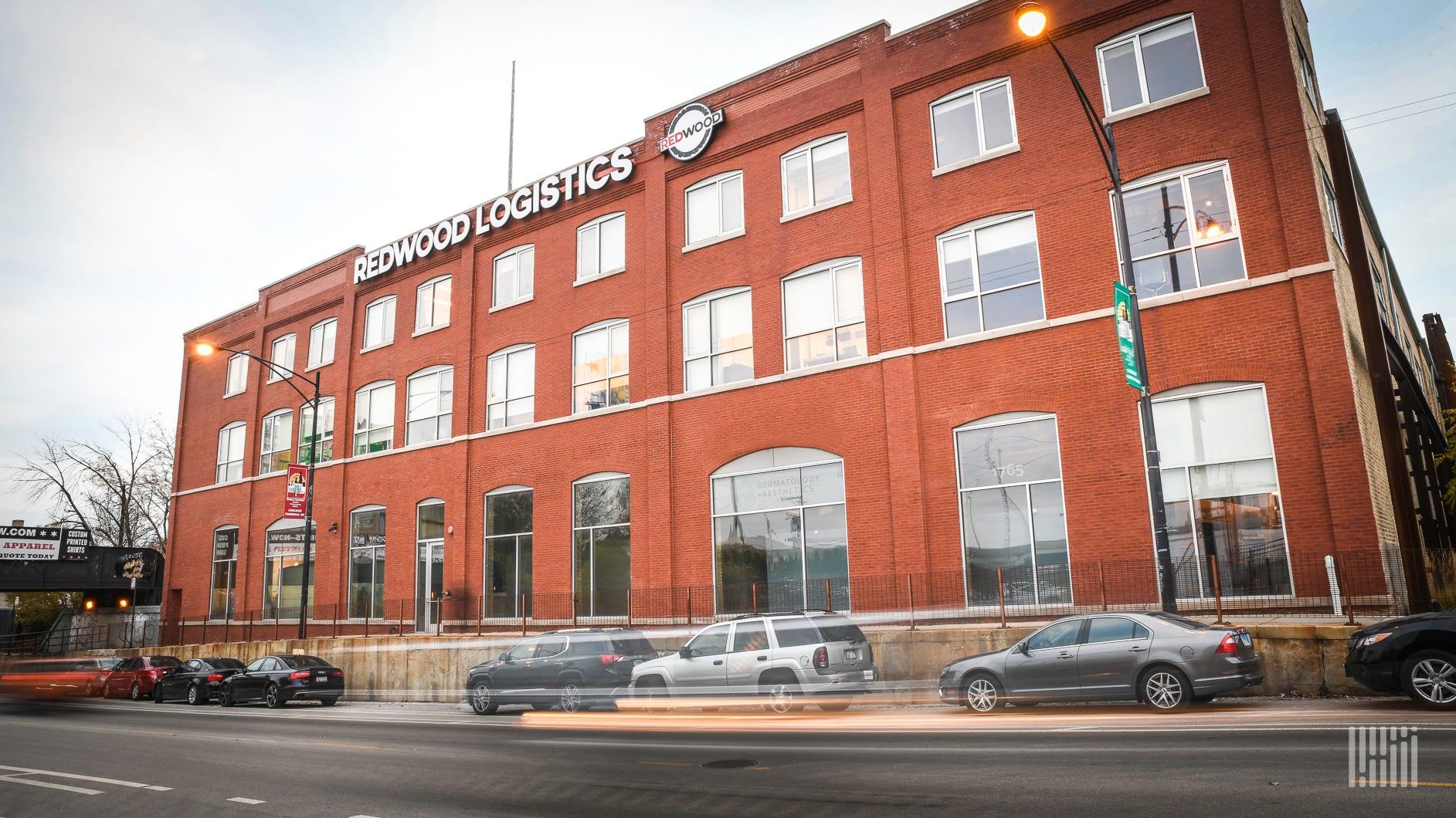 A brick Redwood Logistics building with many windows is shown from across a street.